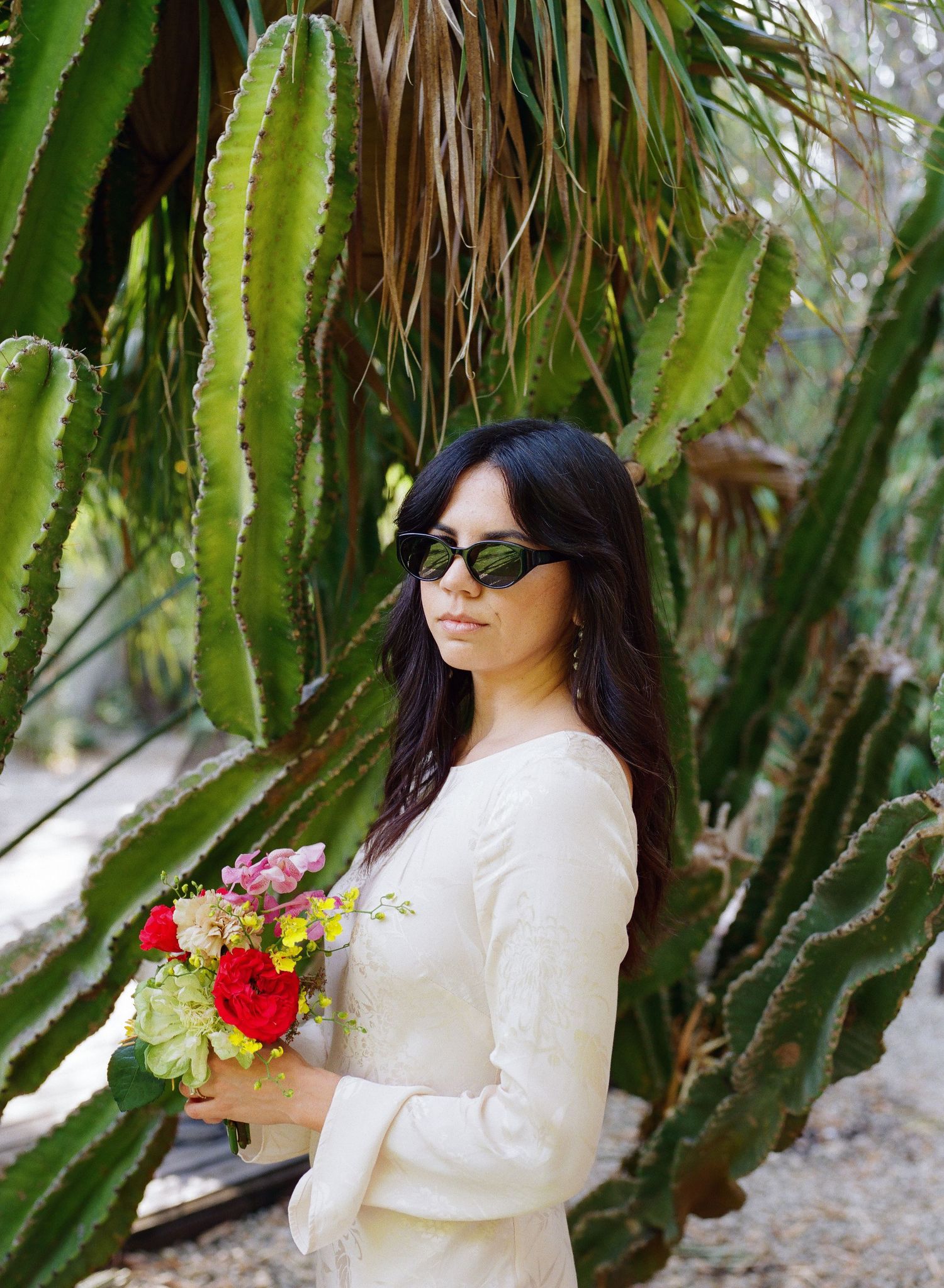Person in white dress and sunglasses posing among tall cactus plants with colorful bouquet.