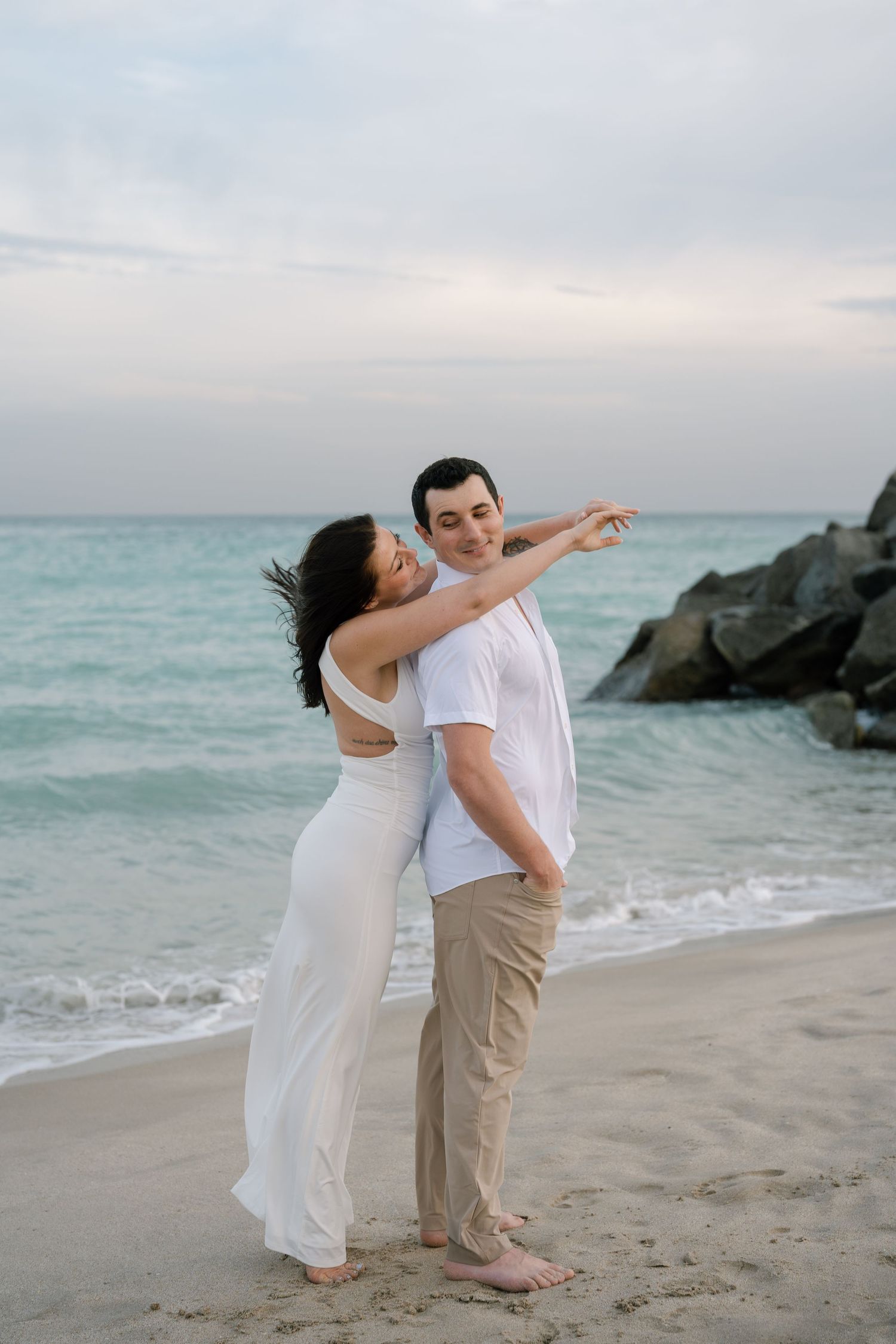 A couple shares fun moments on beach with dramatic rock formations in background.