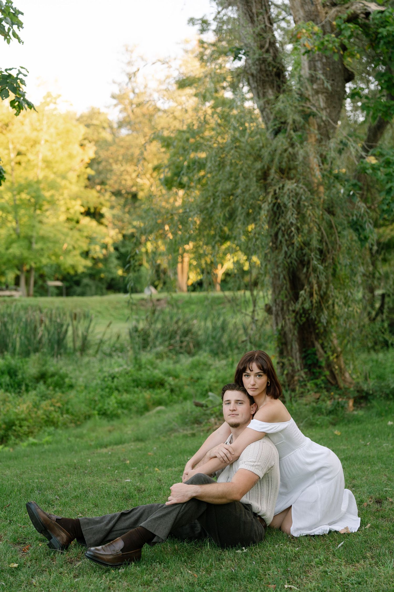 A couple sits together on lush green grass beneath a weeping tree, the woman in a white dress embracing the man from behind.