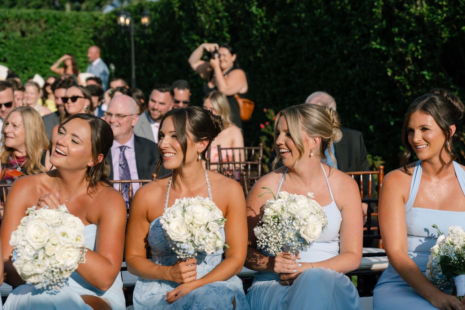 Four bridesmaids in light blue dresses holding white bouquets laughing during outdoor wedding ceremony.