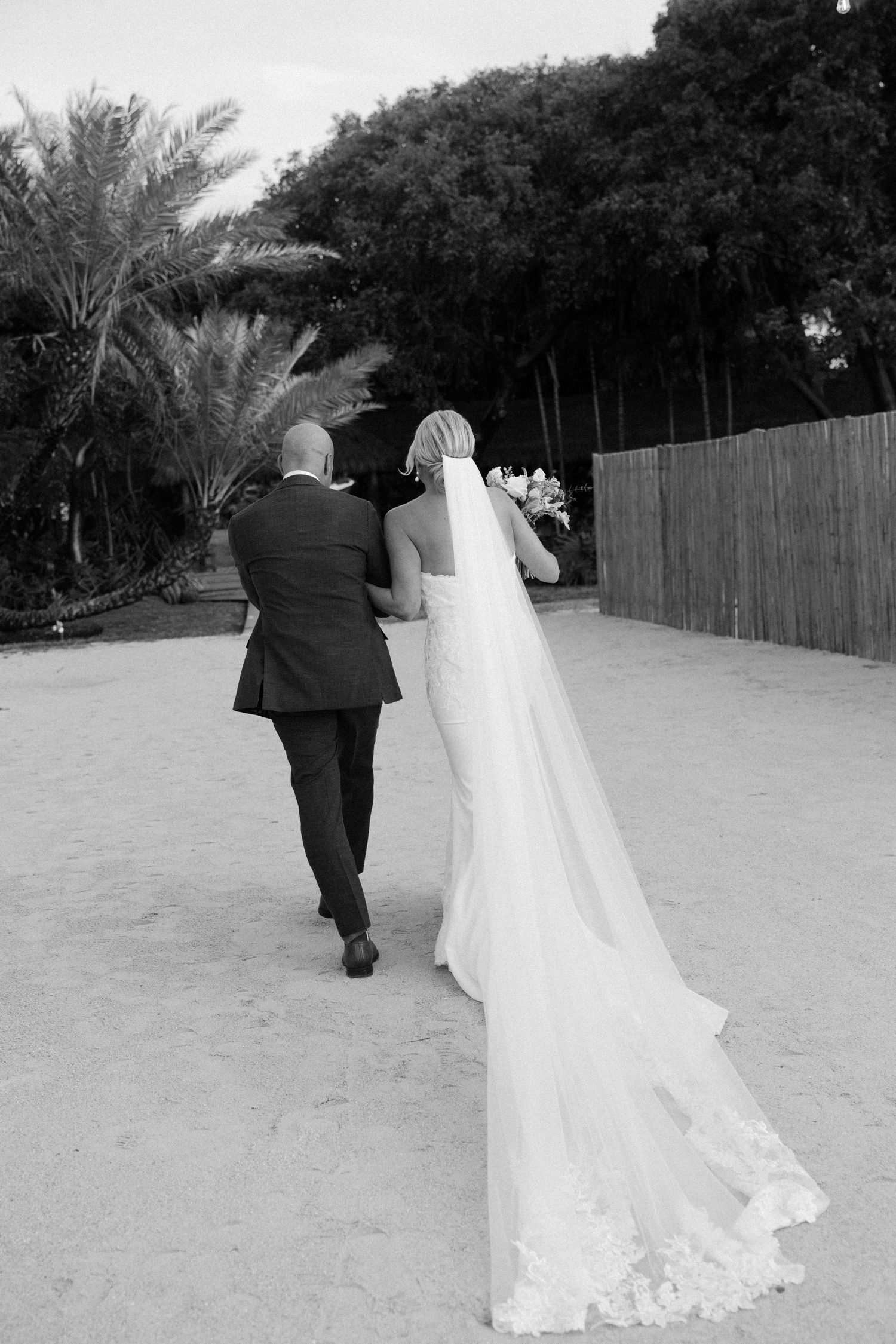 A couple walking away from camera on a sandy beach path during their wedding.