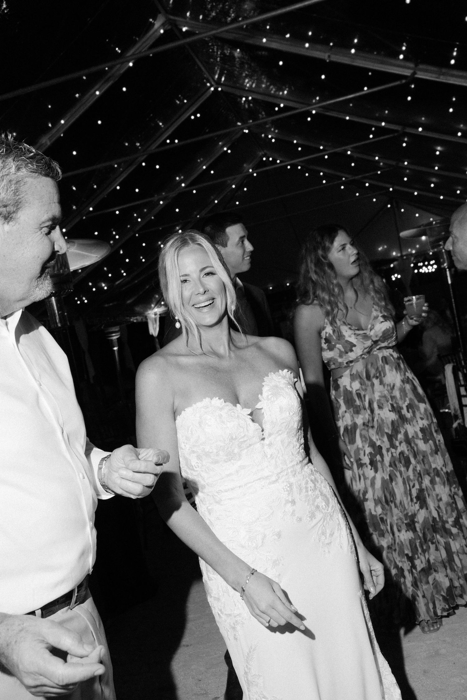 Wedding reception party under twinkling lights with guests dancing and celebrating.