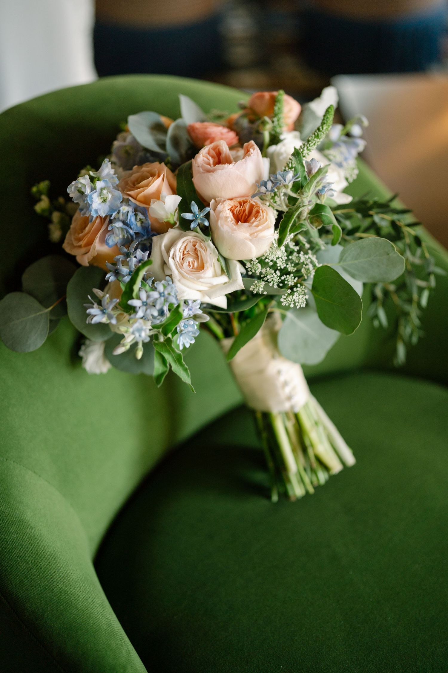 Delicate pastel bouquet with peach roses and blue flowers rests against a green background.