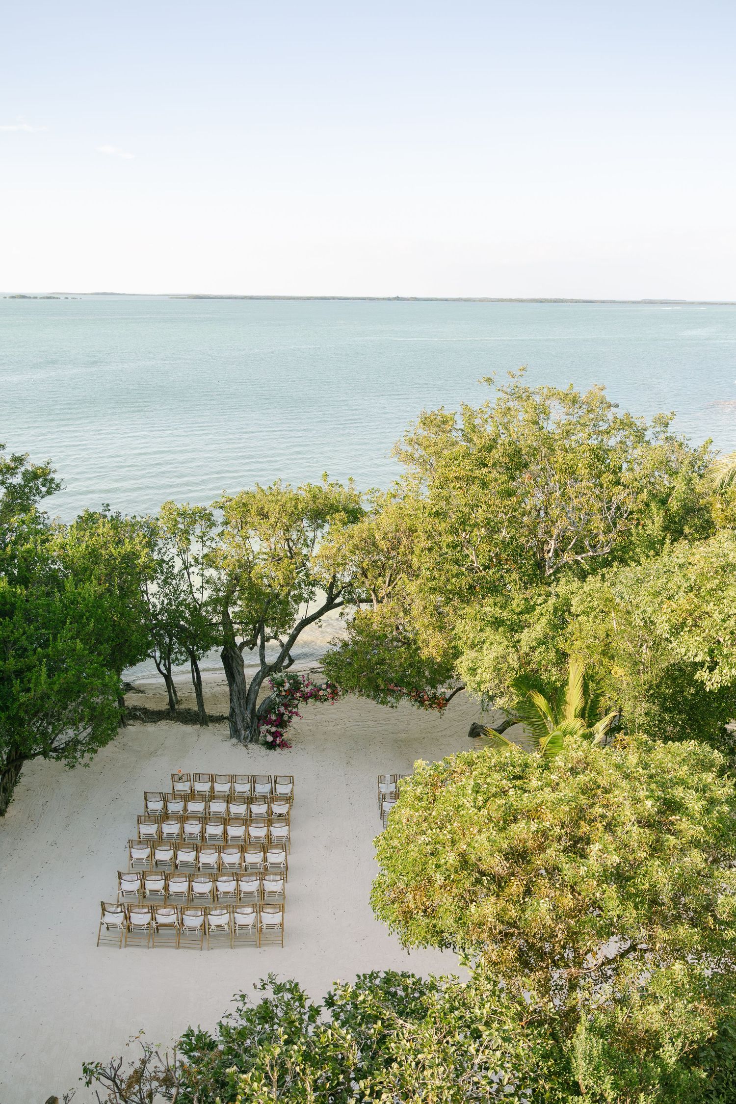 Aerial view of wooden chairs set up for a beach wedding ceremony surrounded by lush trees along the oceanfront.