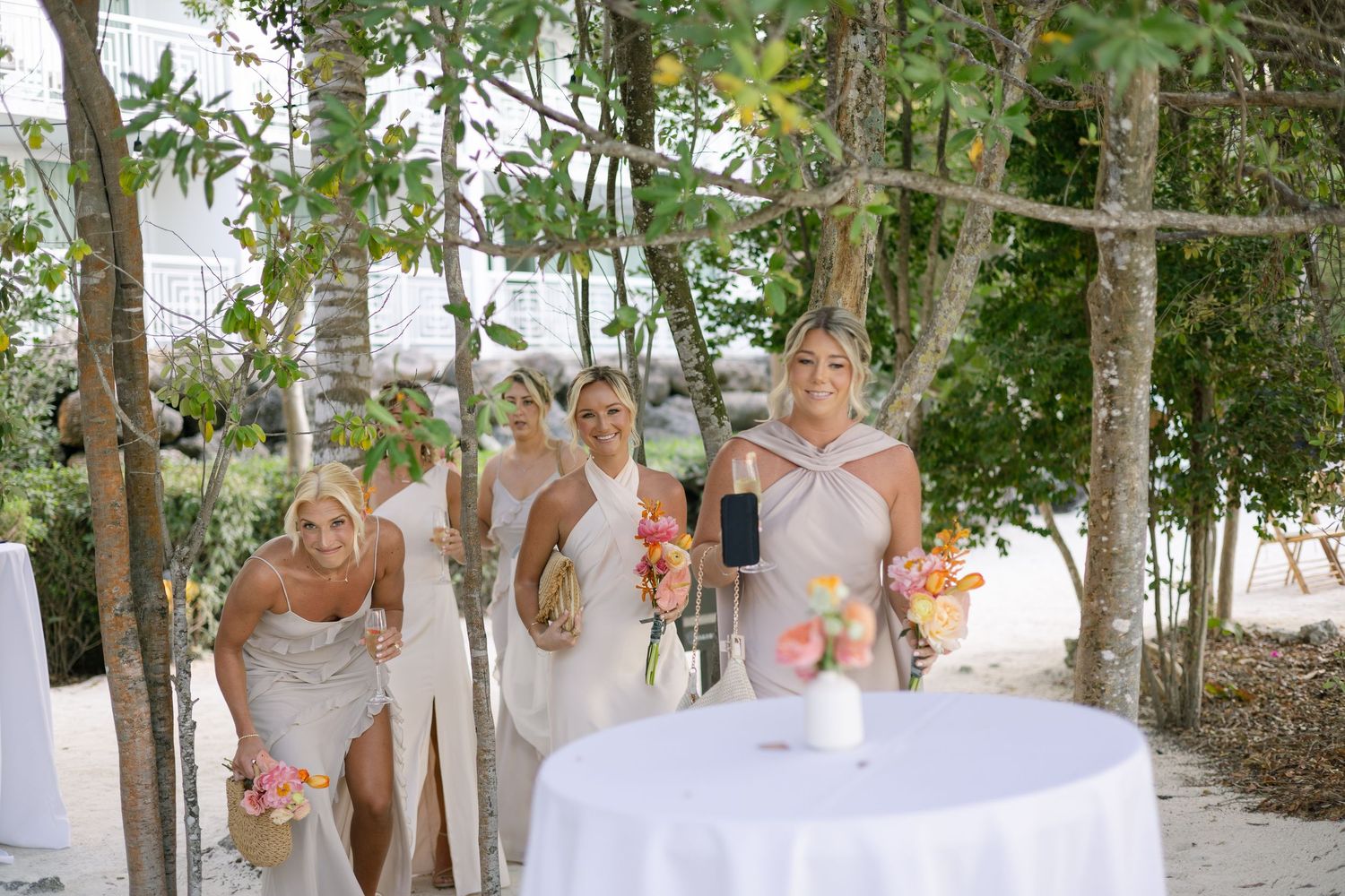 A bridal party in white dresses poses among tropical trees and greenery.