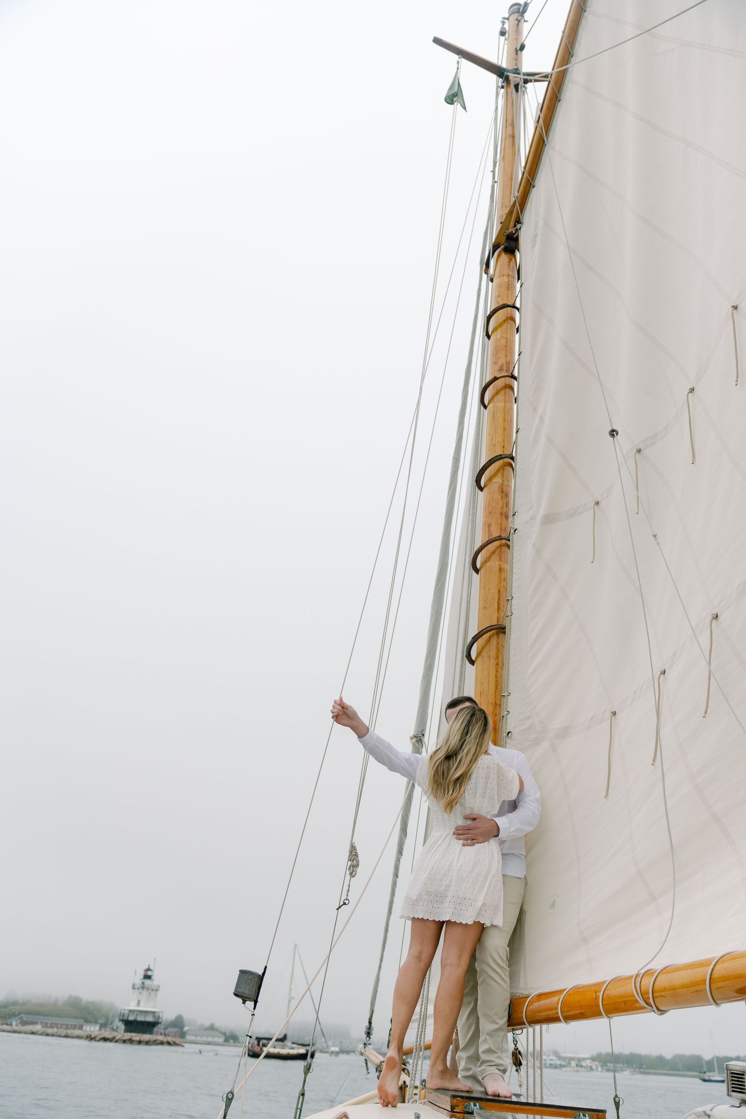 Couple embracing on a classic sailboat deck with white sails and a lighthouse in the misty harbor background.