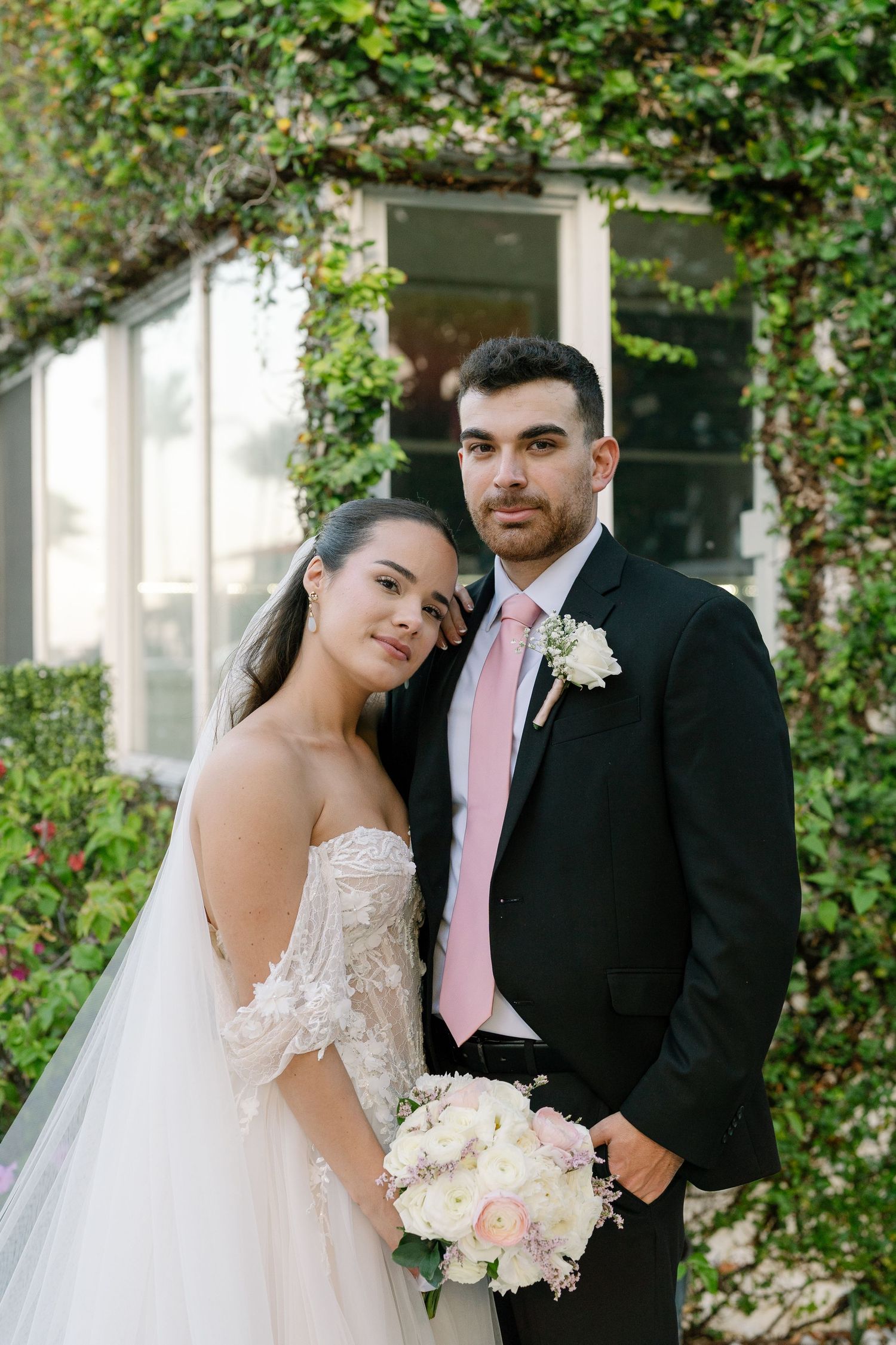 Bride and groom pose together outdoors, she in lace off-shoulder gown, he in black suit with pink tie.