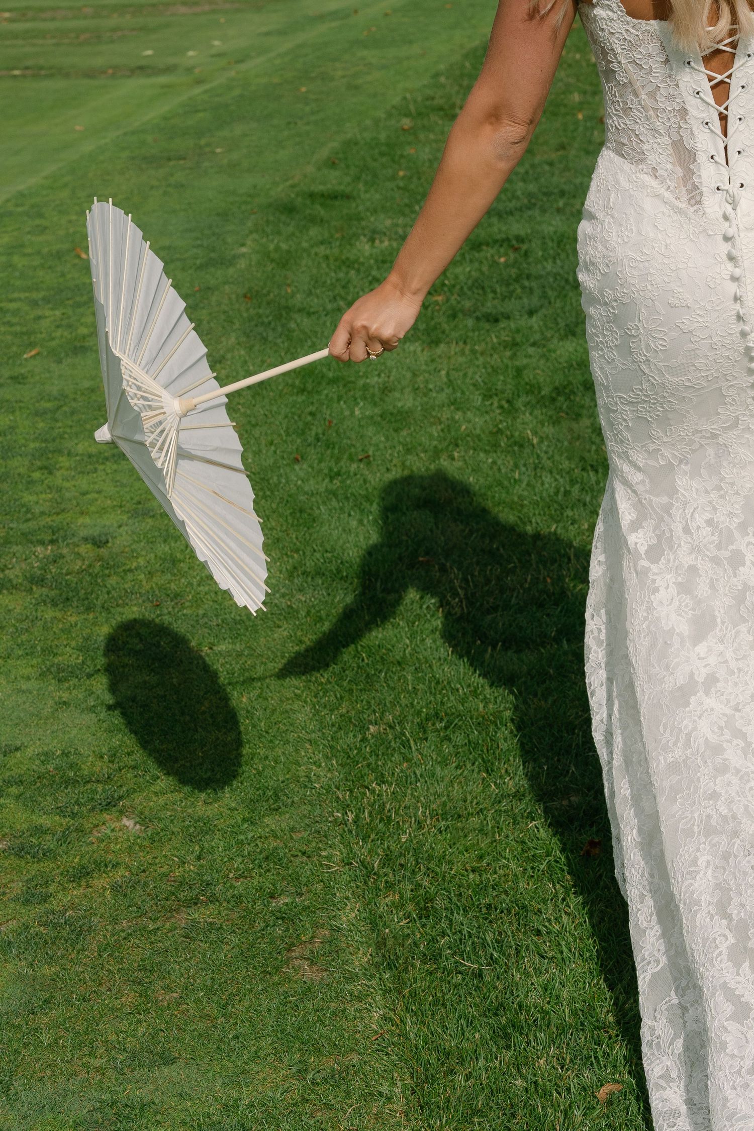 Bride in lace wedding gown holds a white parasol casting a shadow on lush green grass.