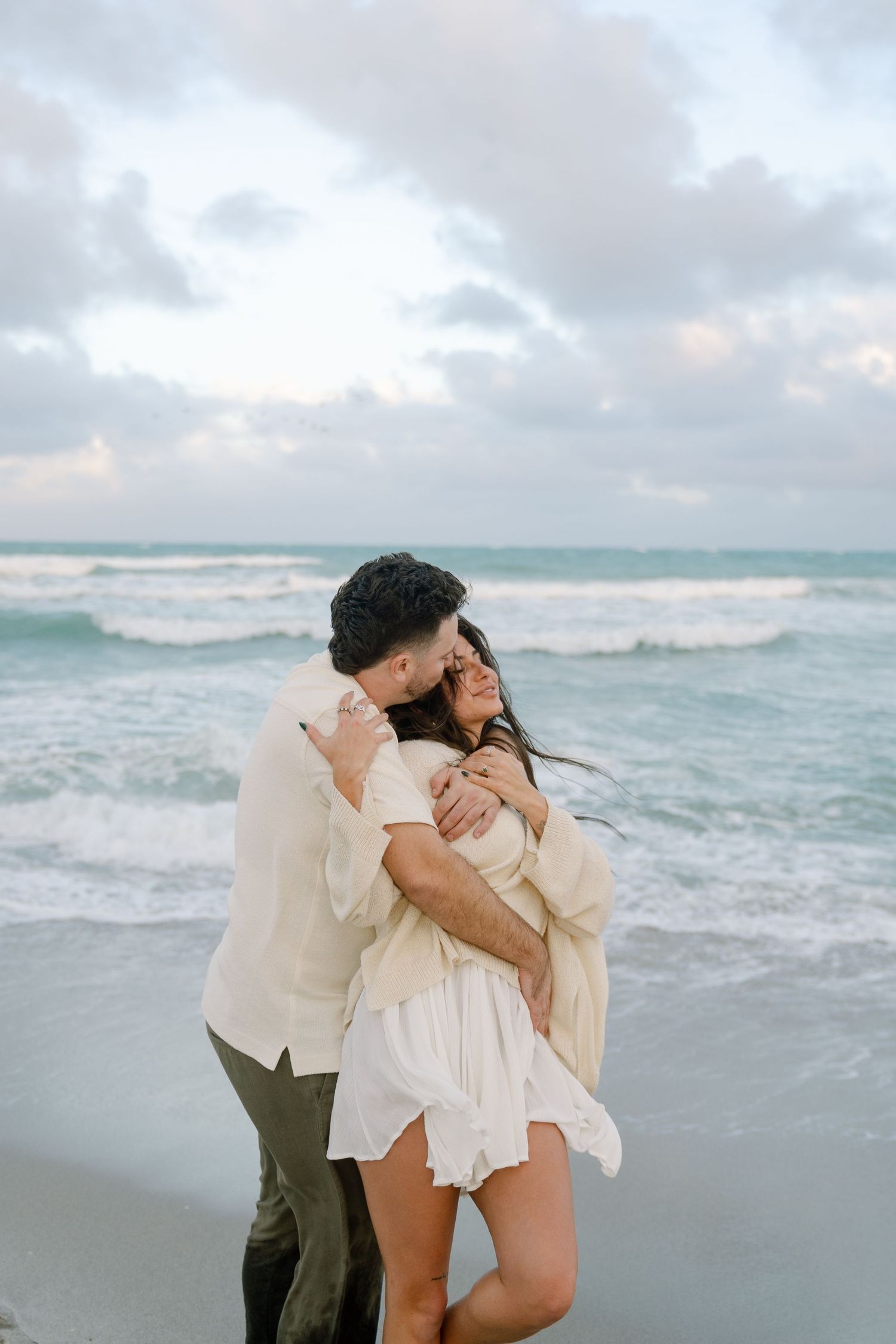 A couple shares intimate moments on a cloudy beach with crashing waves in the background.