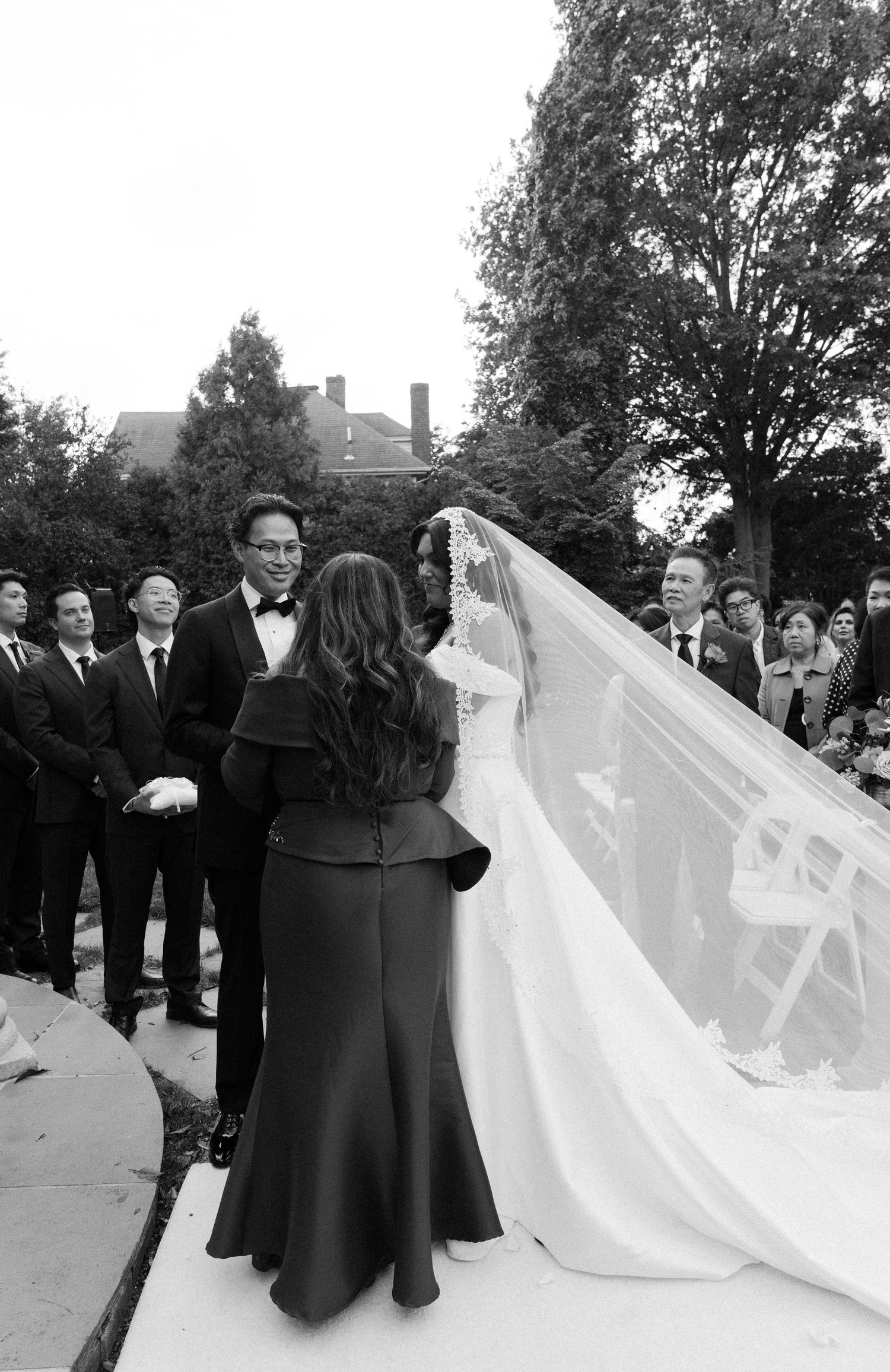 A wedding ceremony scene with guests watching as the bride's veil is adjusted outdoors.