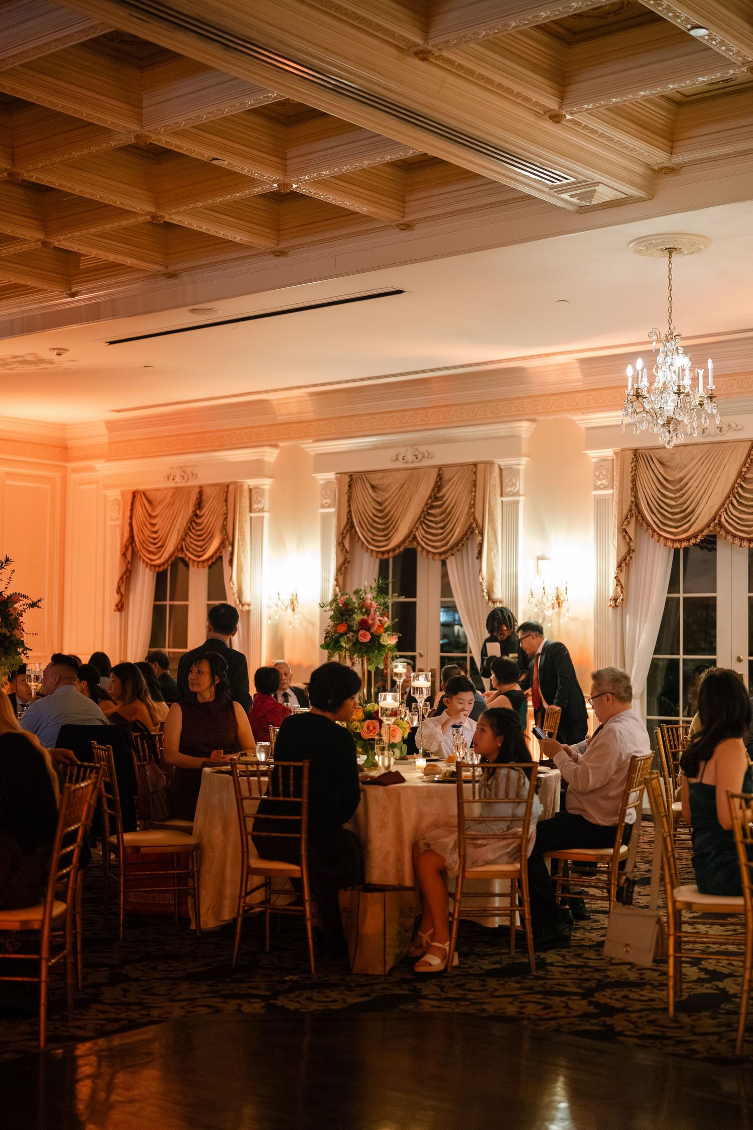 An elegant ballroom reception venue with warm lighting, crystal chandeliers, and draped windows creates a romantic atmosphere.