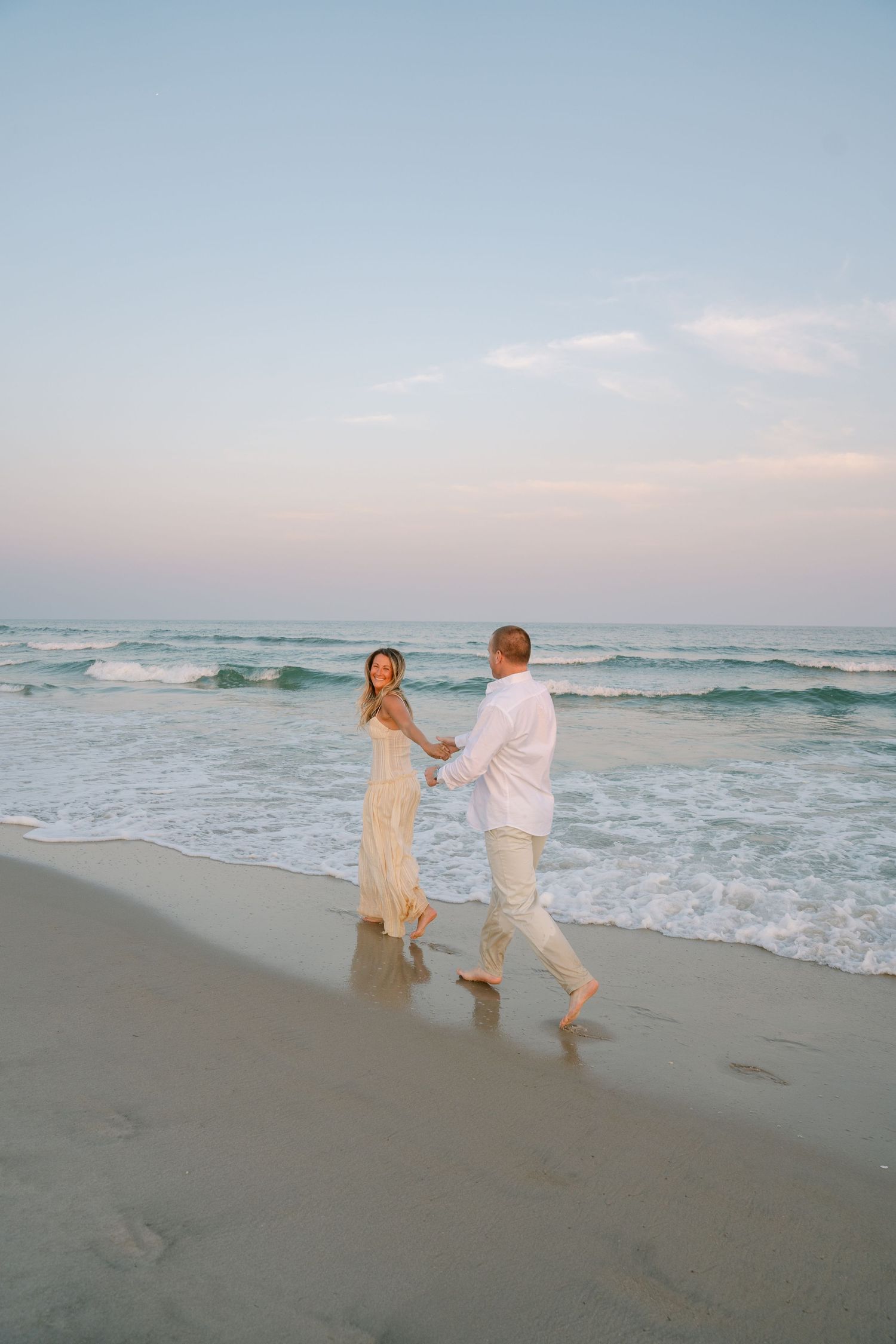 A couple in white outfits dance together on a beach at sunset as ocean waves roll in behind them.