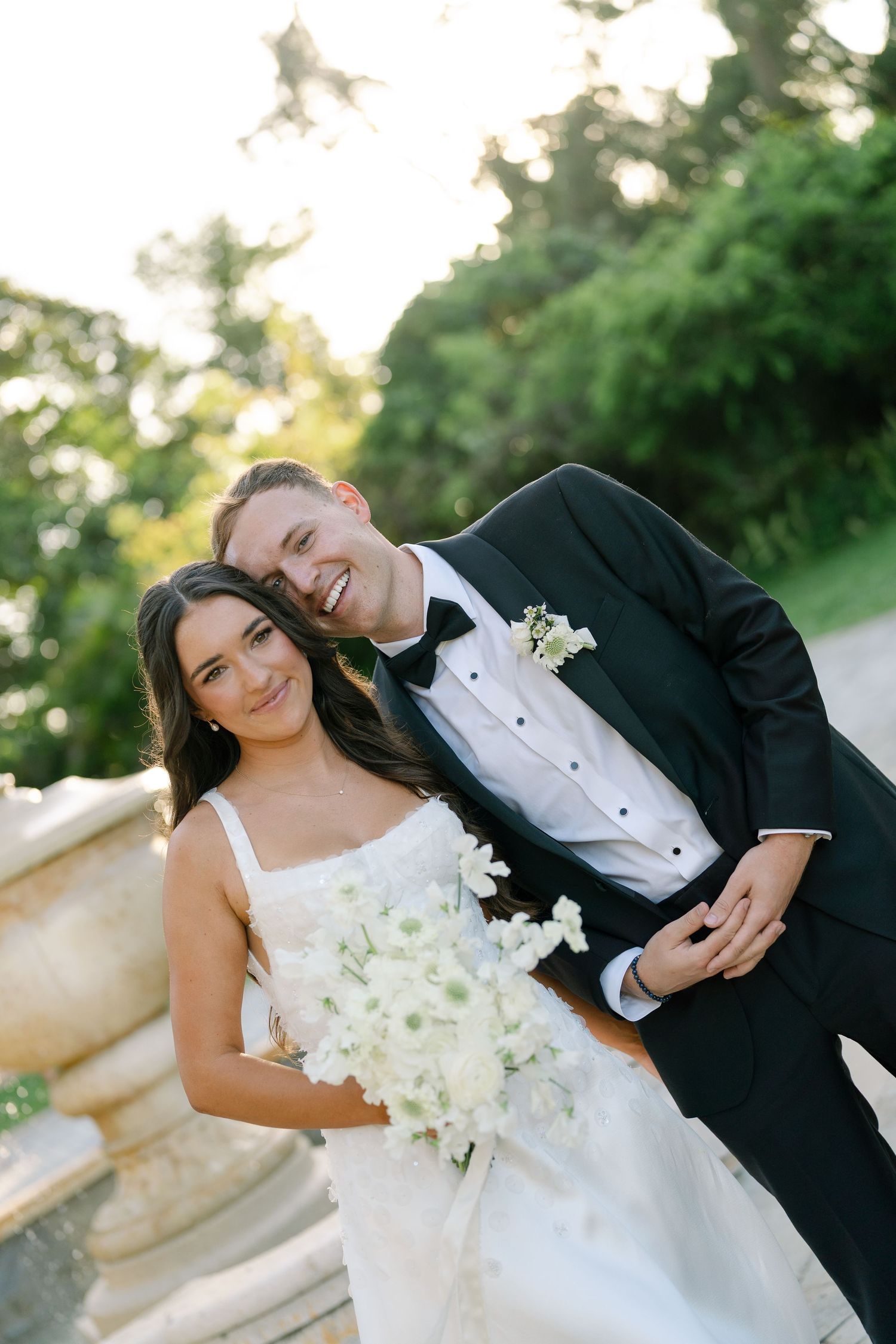 Smiling bride and groom pose together outdoors, bride holding white bouquet, groom in black tuxedo.