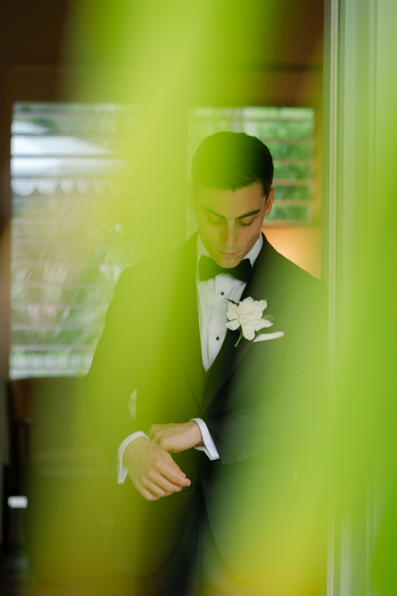 Dapper groom in black tuxedo with white boutonniere adjusts cufflinks, photographed through lush green foliage.