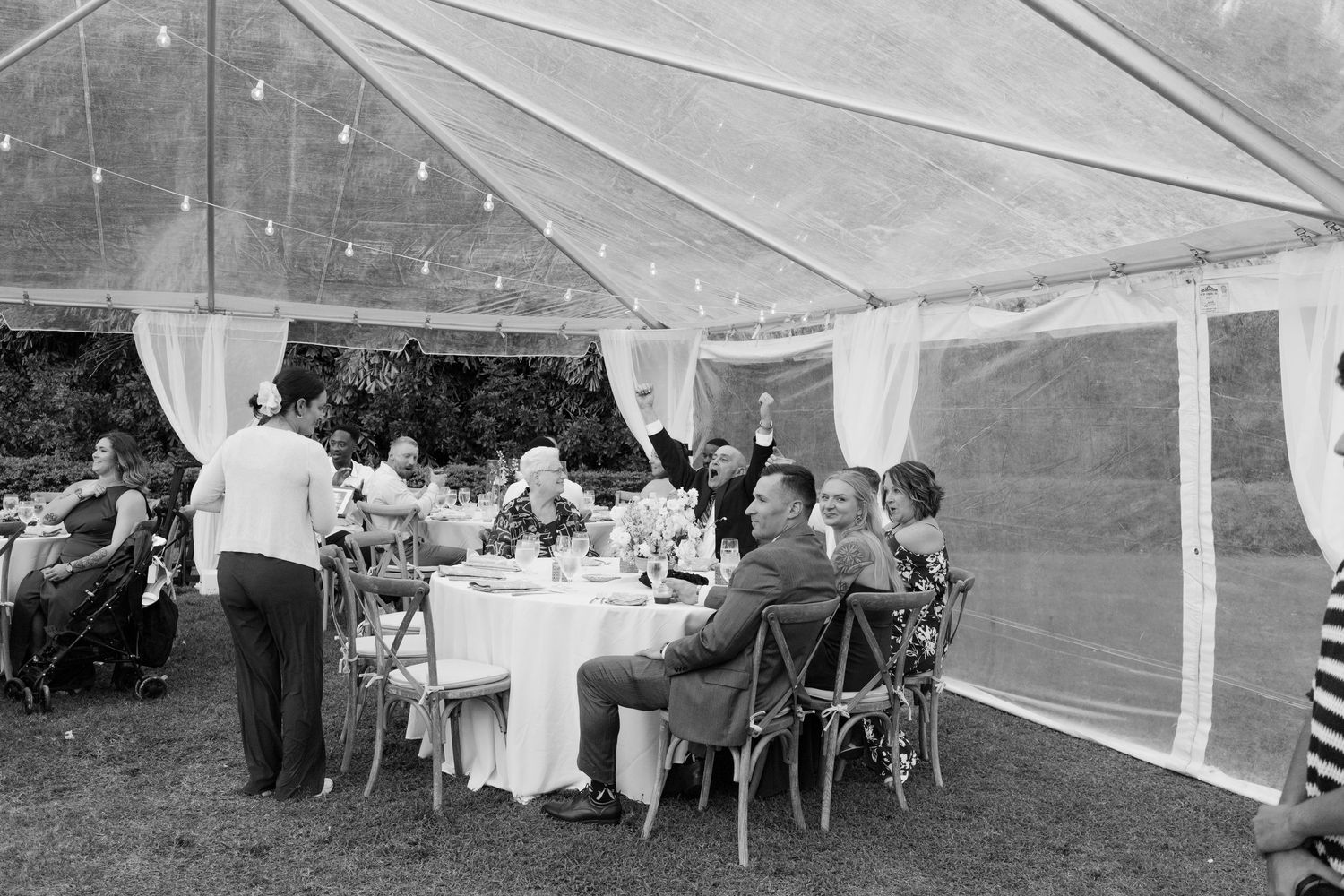 Black and white photo of wedding guests dining and celebrating under a clear tent with string lights.