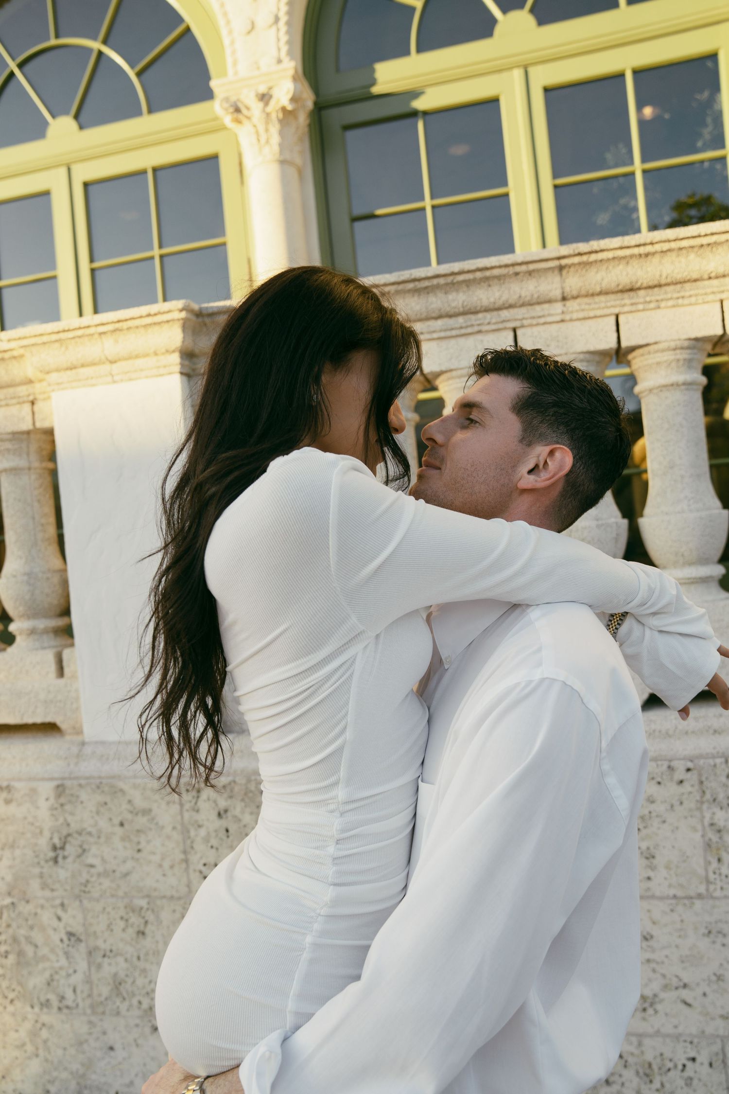 Romantic couple in all white outfits share a tender moment against an elegant architectural backdrop with marble pillars.