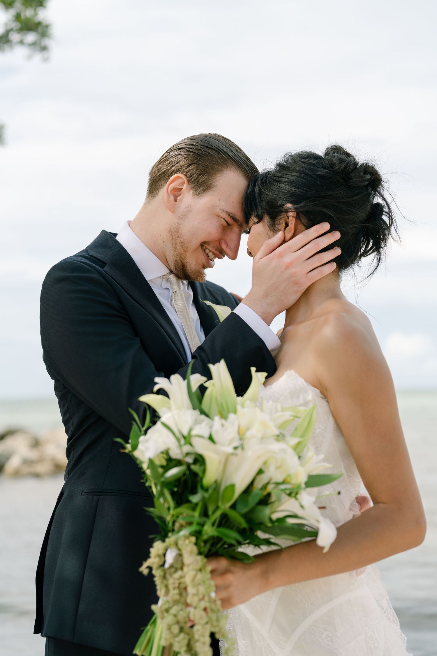 A groom in a black suit tenderly holds his bride's face as they touch foreheads, she holds white lily bouquet by the water.