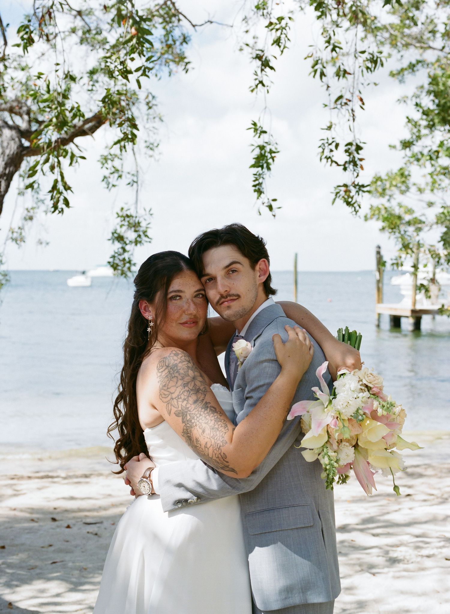 Tattooed bride and groom embrace on sandy beach with ocean backdrop holding a pink and cream bouquet.