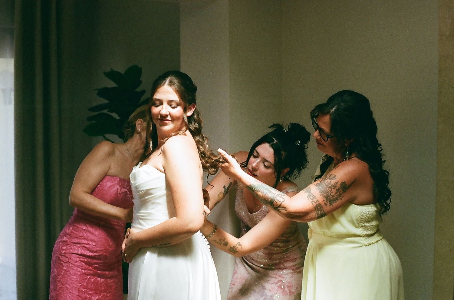 Three women in formal dresses assist each other with their hair and makeup.
