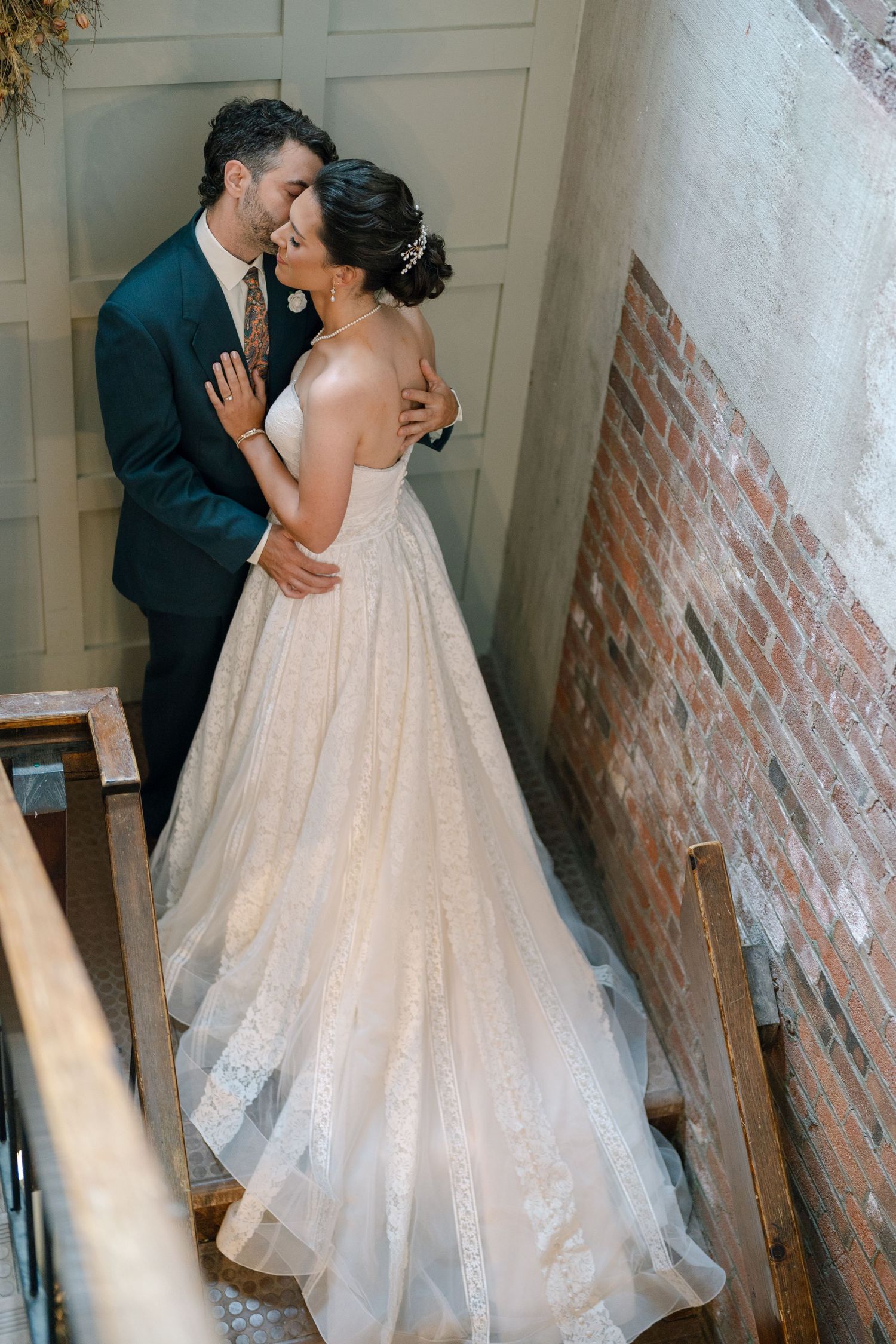 Romantic wedding moment shows a couple in formalwear sharing an intimate kiss against a rustic white and brick wall backdrop.