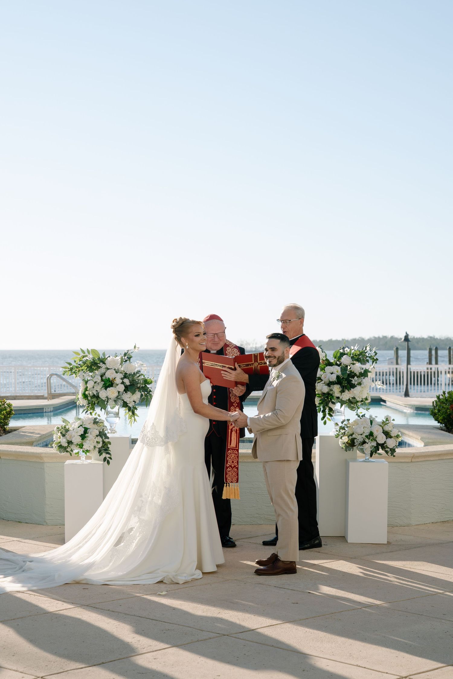 Outdoor waterfront wedding ceremony with white floral arrangements and guests in attendance.