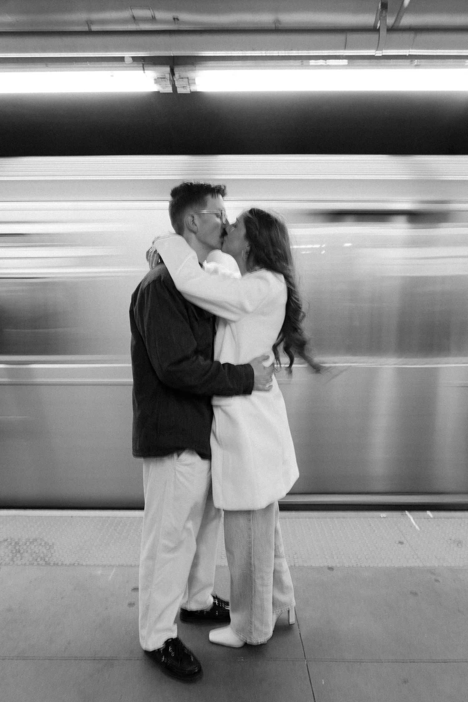 Black and white photo of a couple embracing on a subway platform as trains blur past in motion.