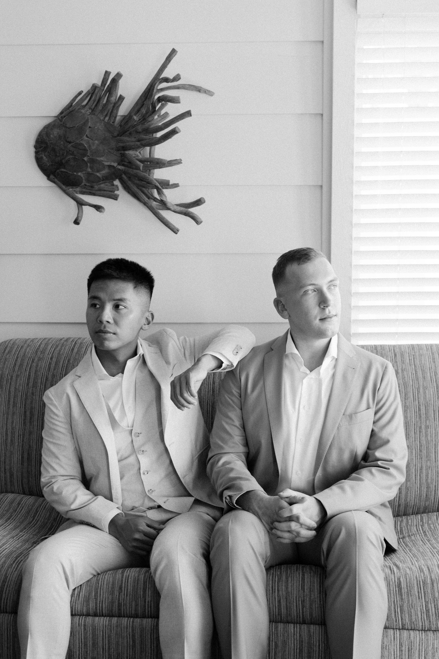 Two people in light suits sit on a gray couch beneath a dark wall decoration.