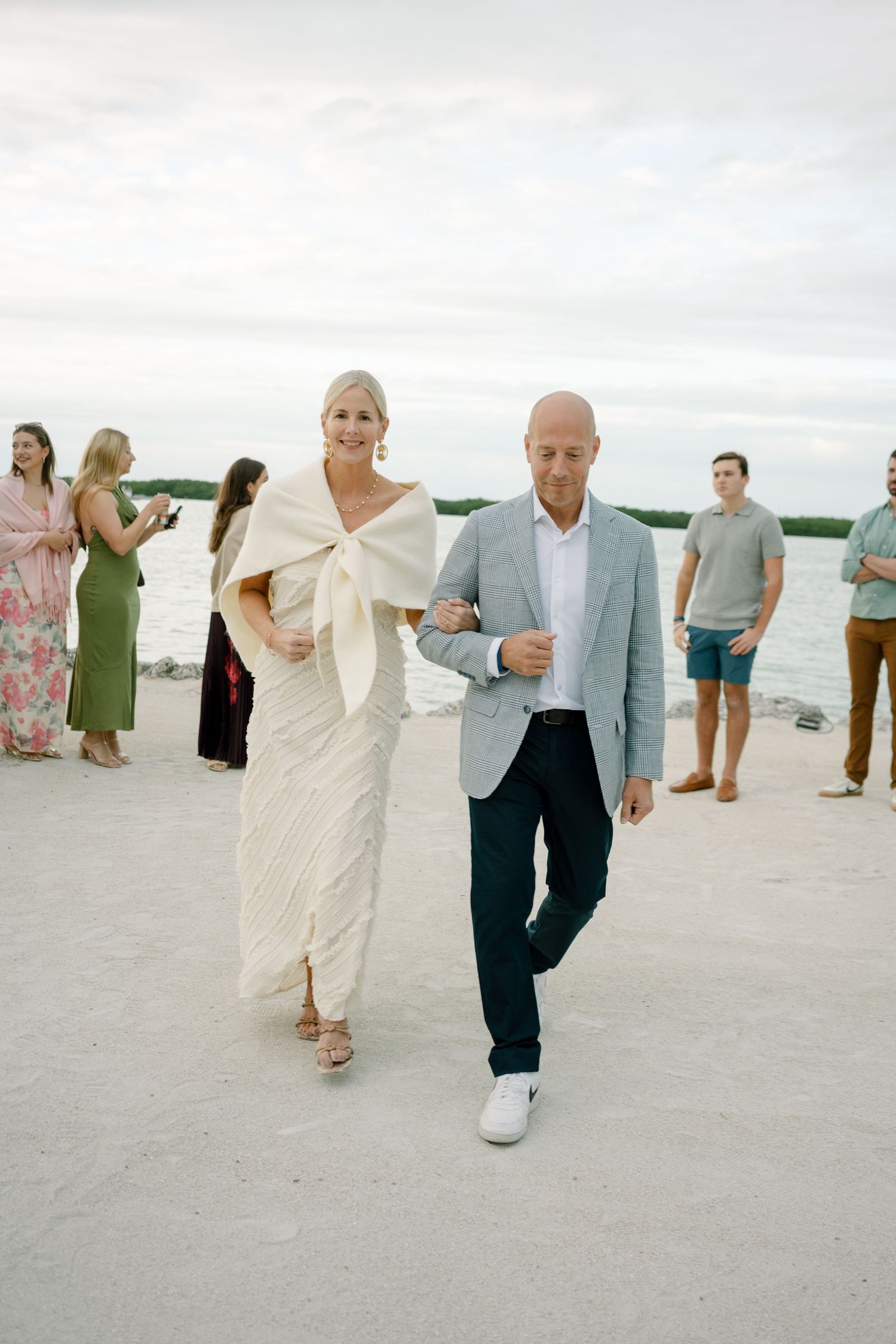 A wedding couple walks arm in arm on a beach with guests in the background on an overcast day.