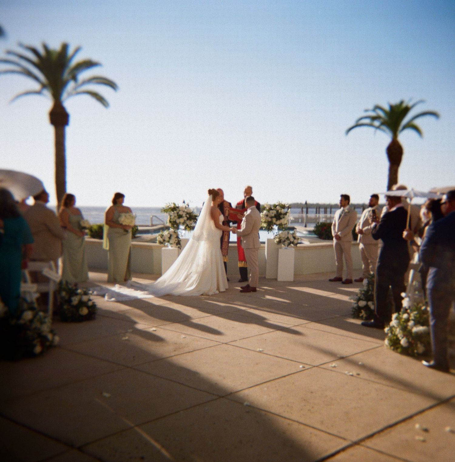 Wedding guests gather on a sunny terrace with palm trees and ocean views during an outdoor ceremony.