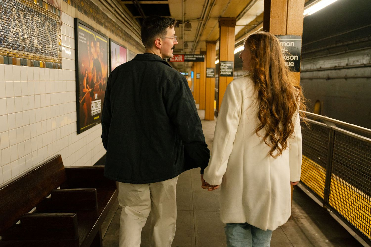 A couple walks together on a subway platform with tiled walls visible in the background.