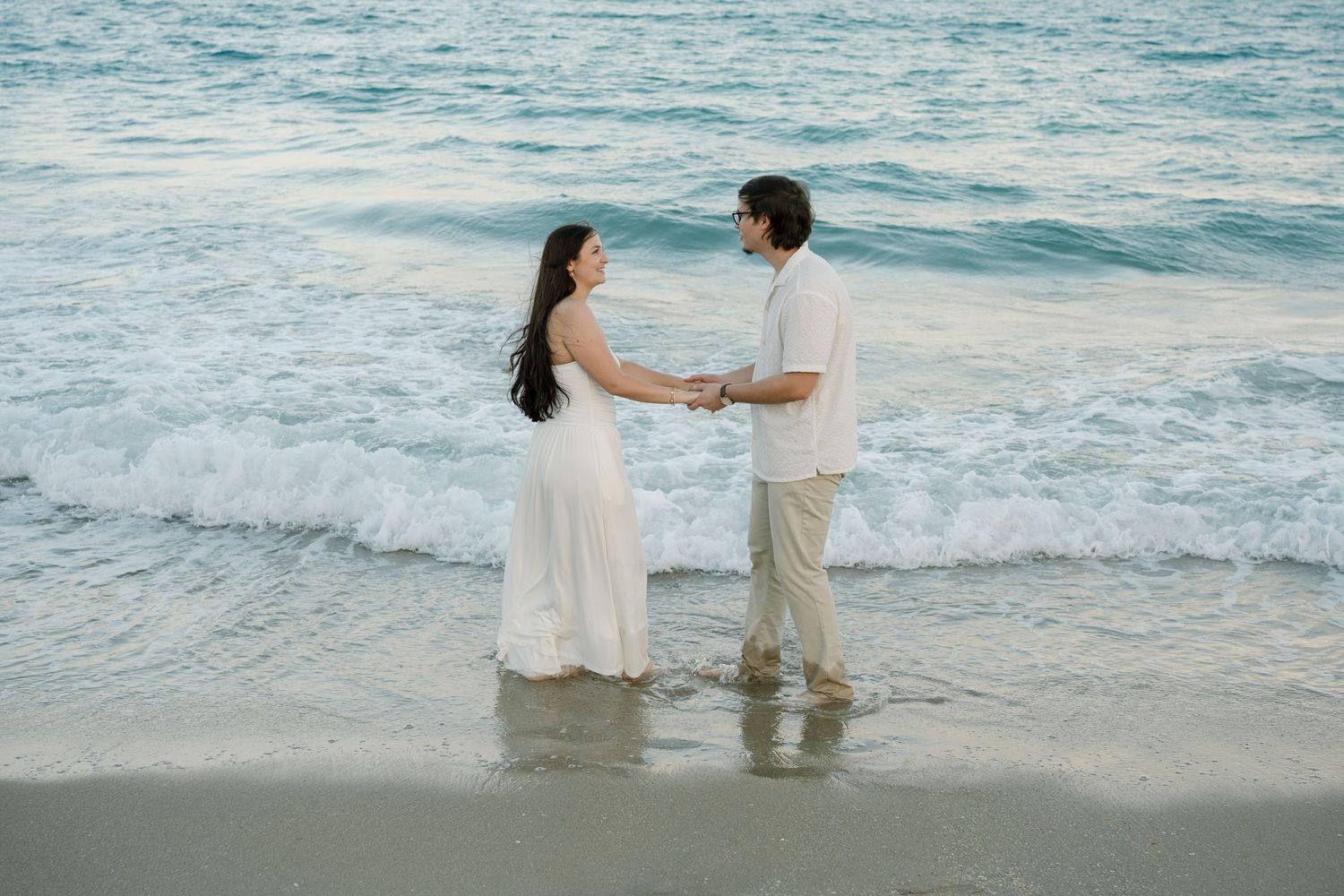 A couple holds hands and dances together at the edge of the ocean waves on a sandy beach at sunset.