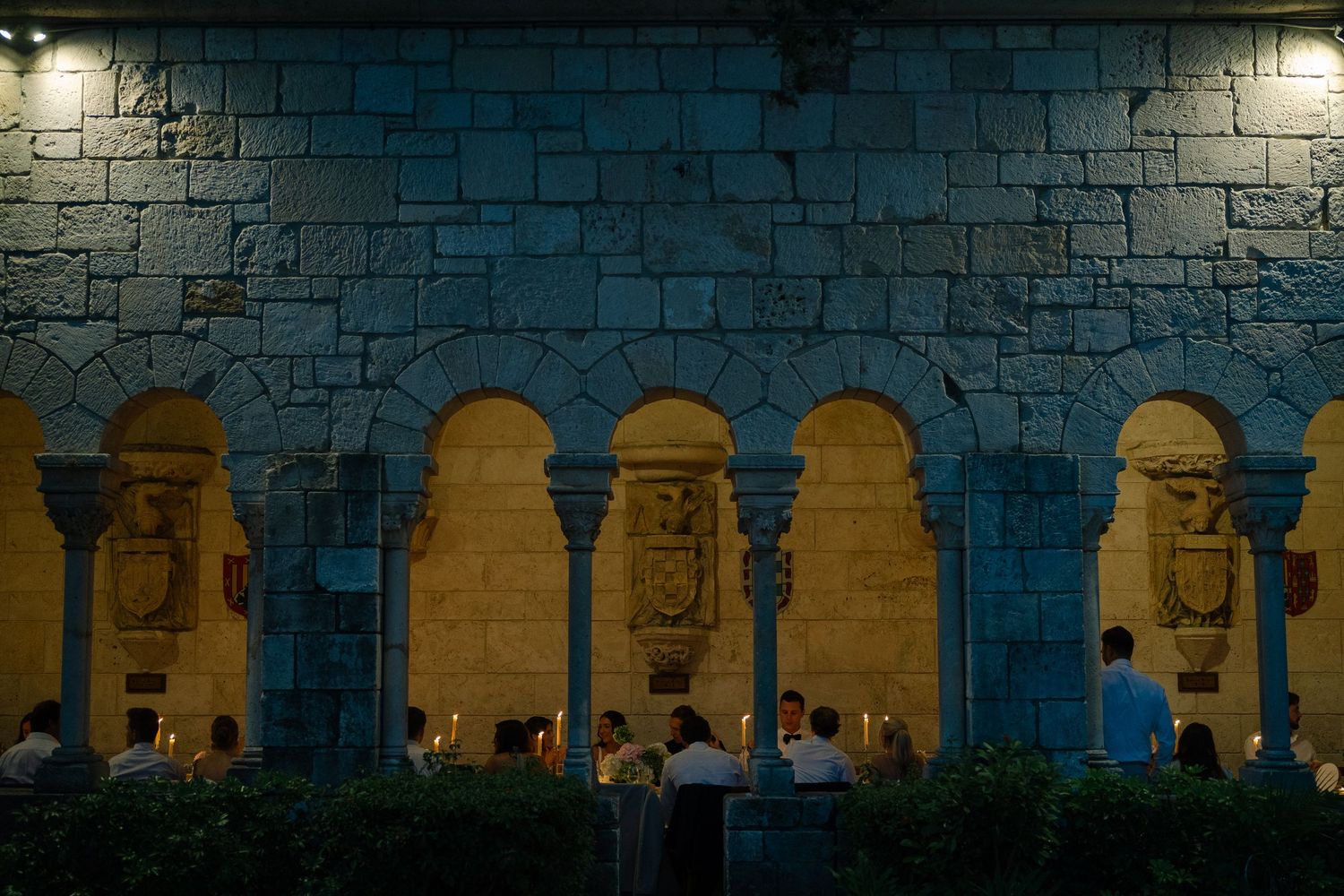 Elegant candlelit wedding reception in a historic stone cloister with arched columns at night.