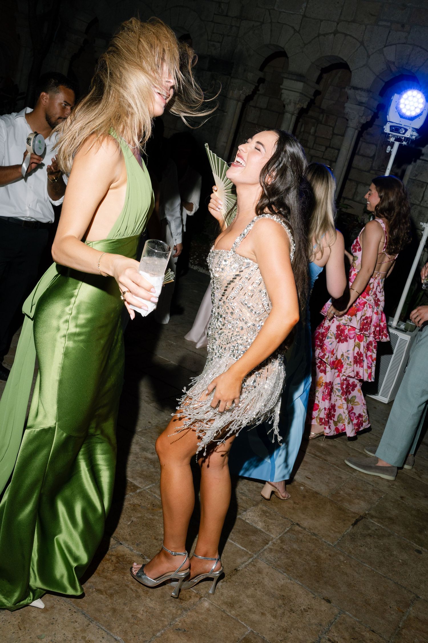 Two women dancing joyfully at wedding reception, one in green satin gown and one in silver fringe dress.