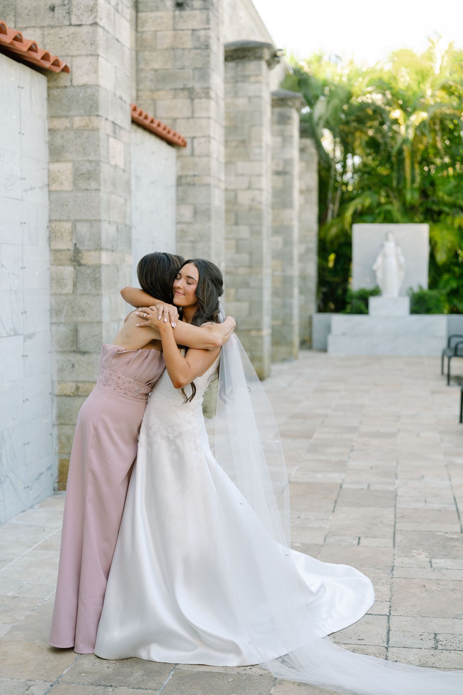 Bride in white gown and veil shares a warm embrace with bridesmaid in pink dress outside stone church.