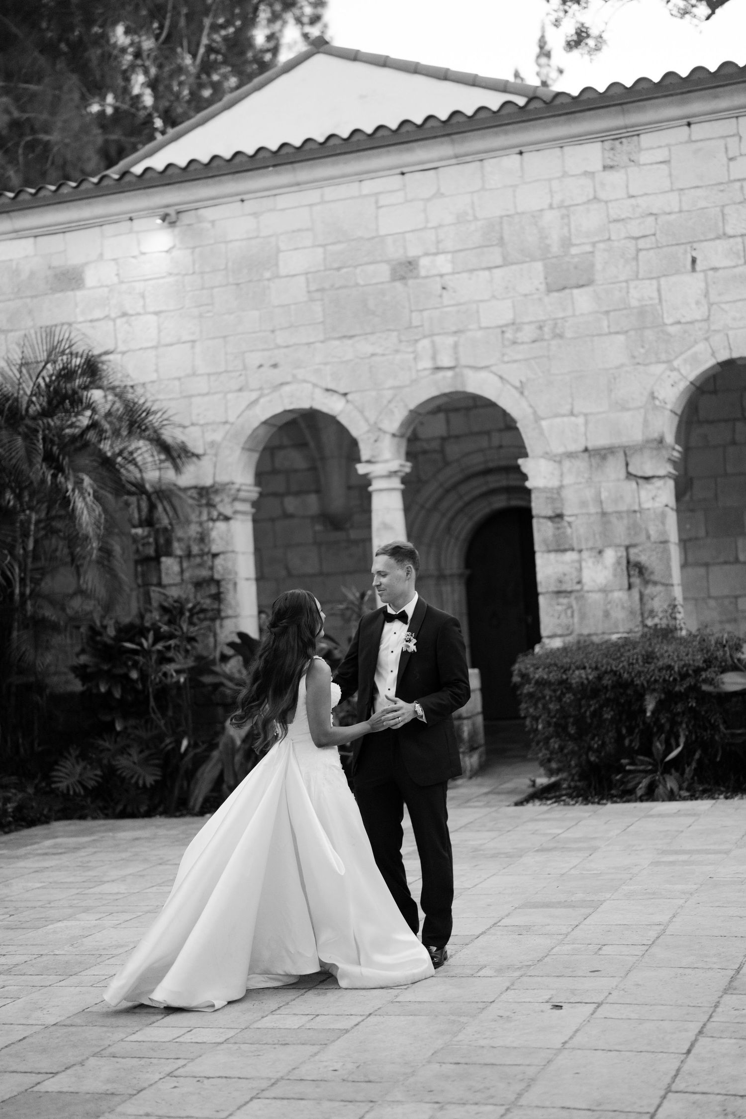 Black and white photo of bride and groom walking together outside historic stone arched wedding venue.
