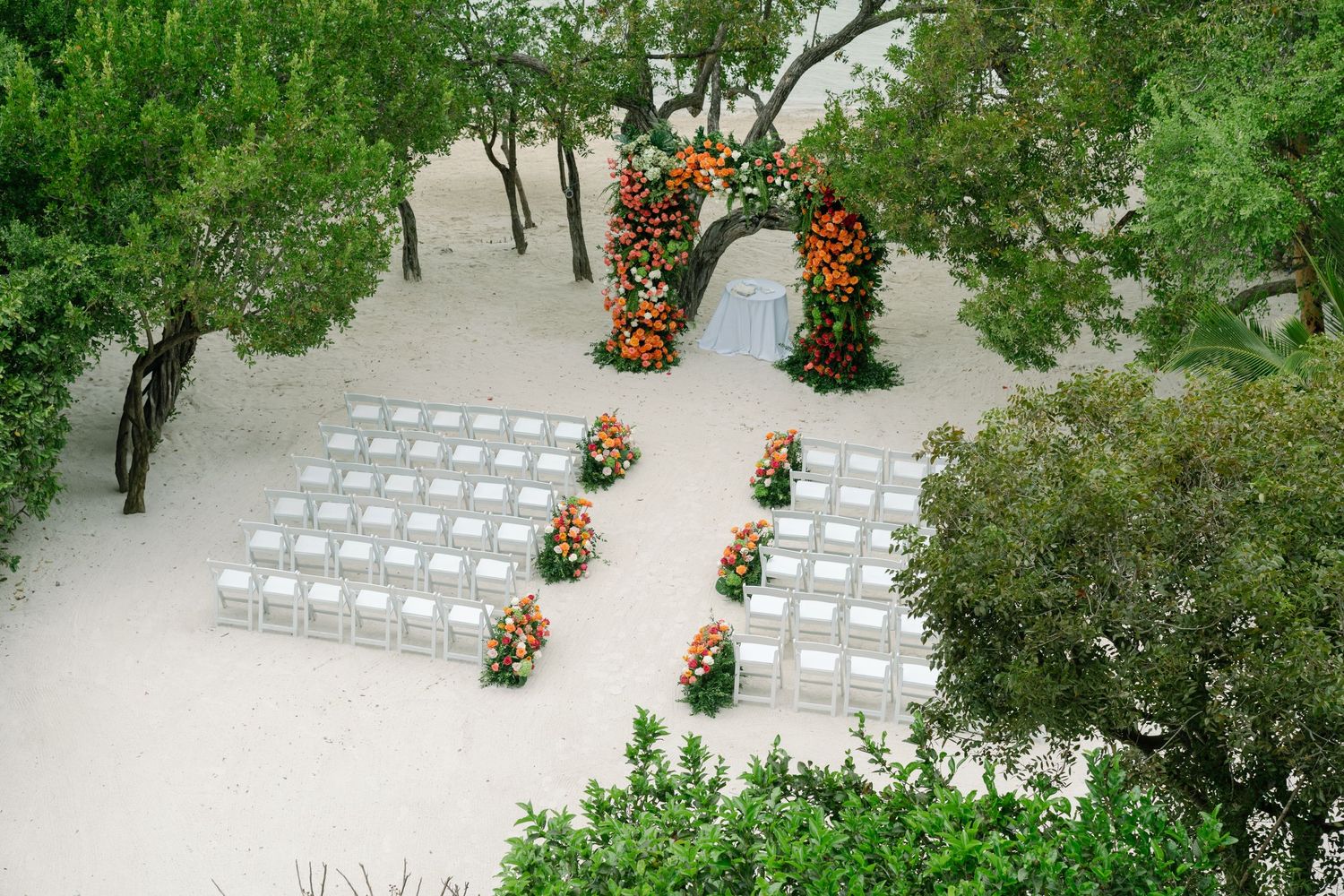 Outdoor wedding ceremony setup with white chairs and floral arch surrounded by lush greenery, viewed from above.