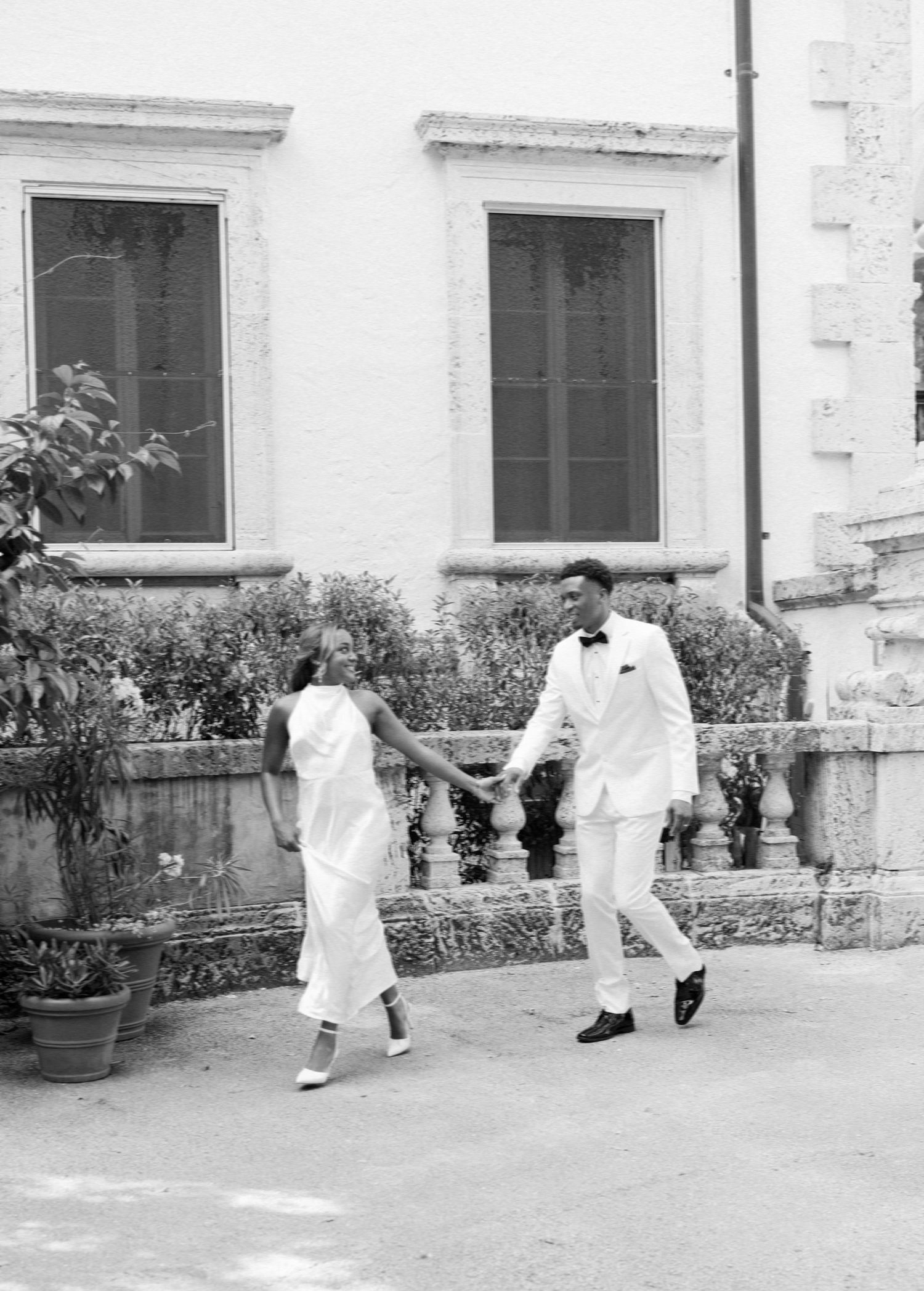 A couple in white formal attire walks together on a path in front of a building with two windows.