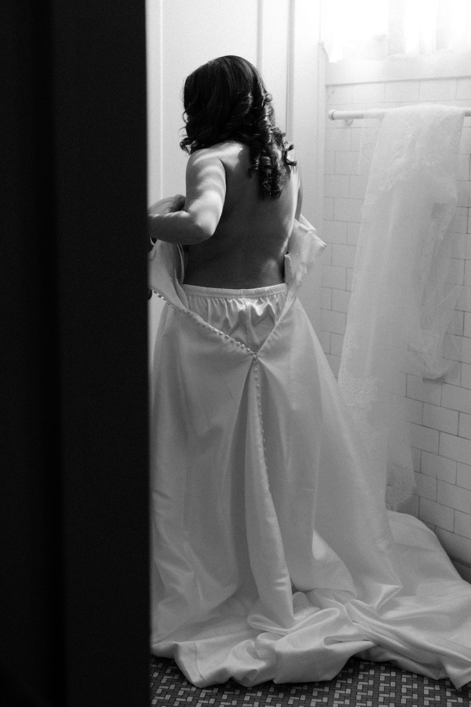 Black and white photo showing back view of someone in a flowing white wedding dress with curled dark hair.