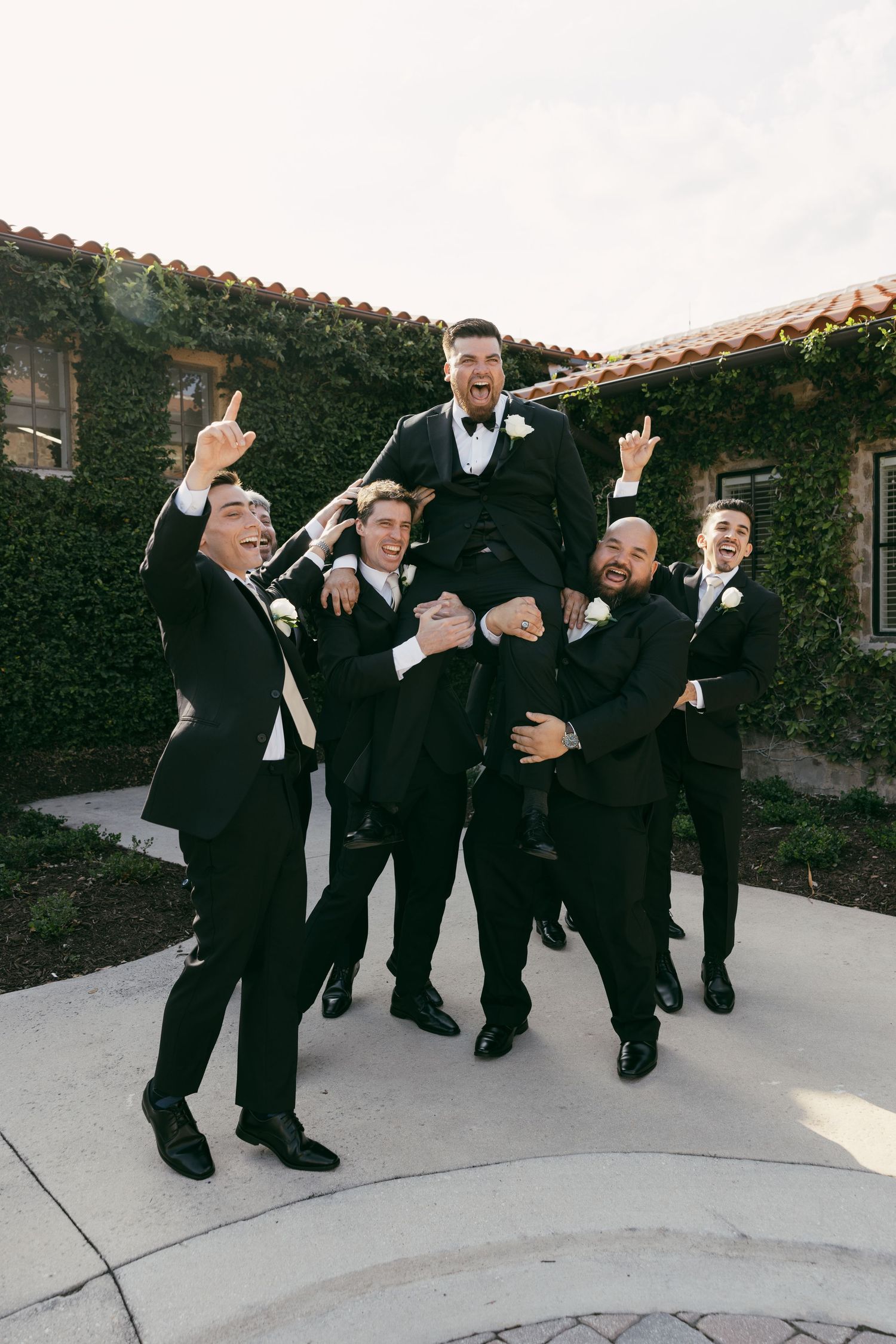 Groomsmen in black tuxedos lifting and celebrating with playful poses outdoors.
