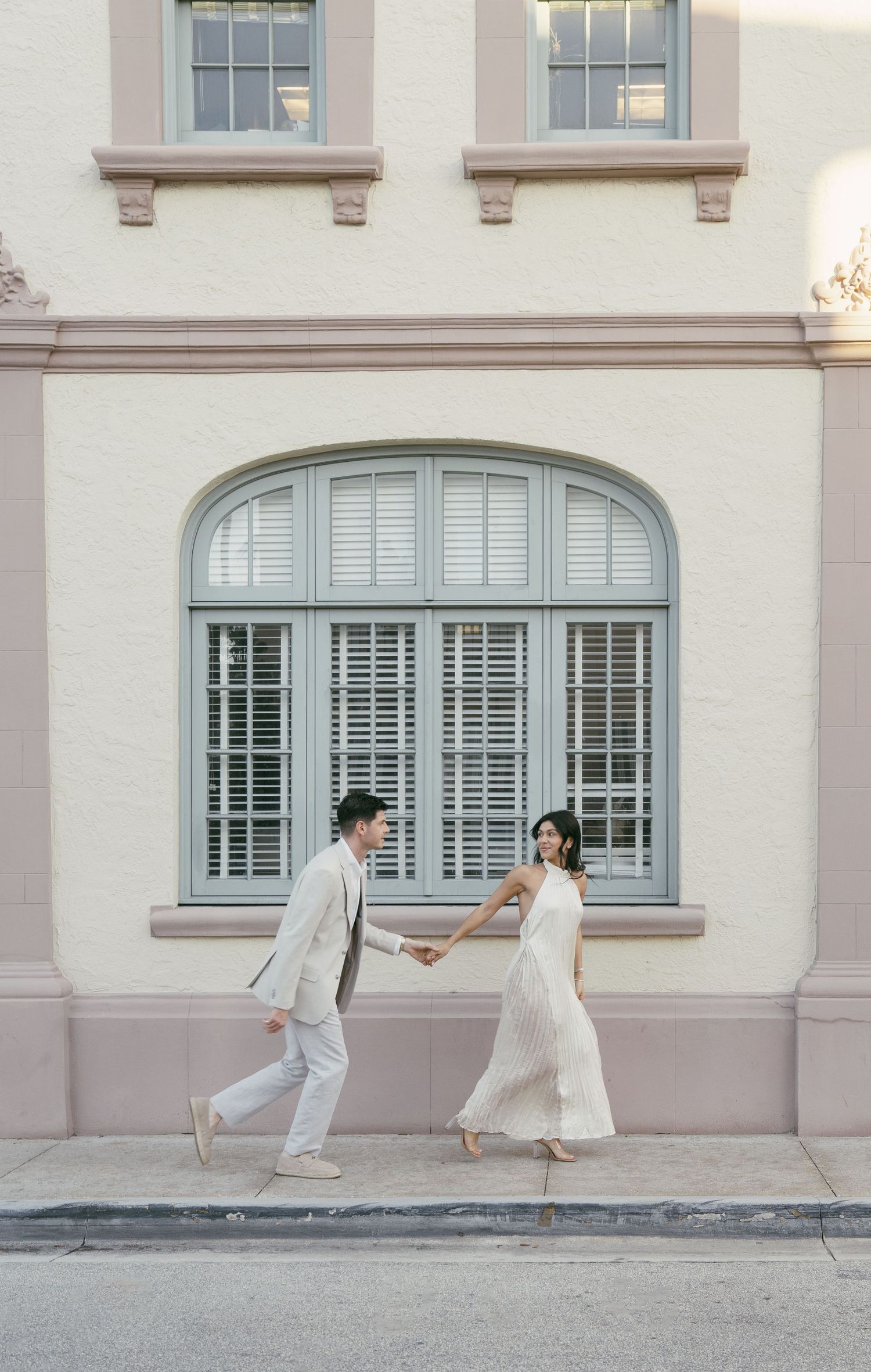 A couple in light-colored wedding attire dancing playfully in front of an elegant arched window with shutters.