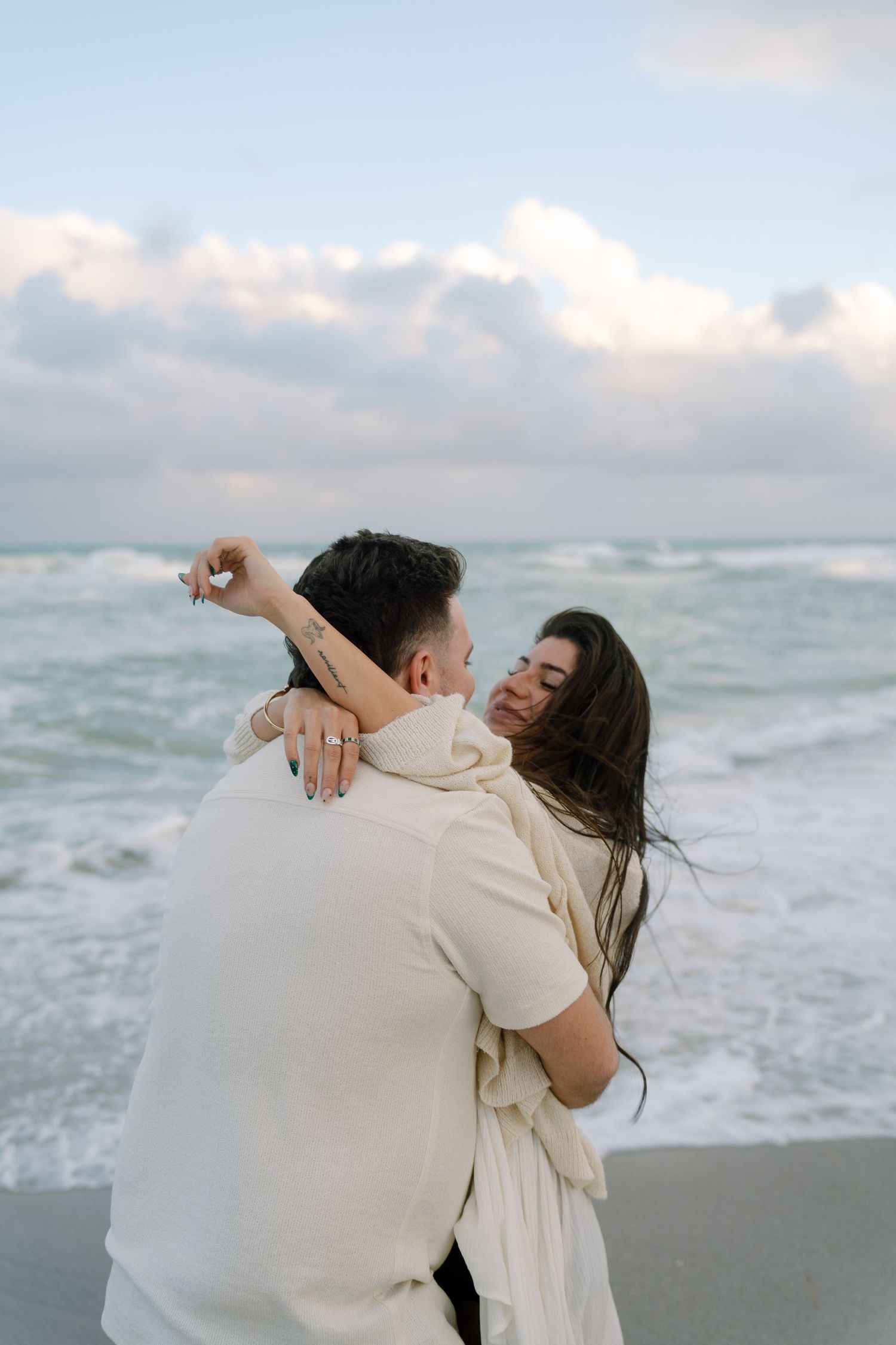 A couple embraces lovingly on a cloudy beach with crashing waves in the background.
