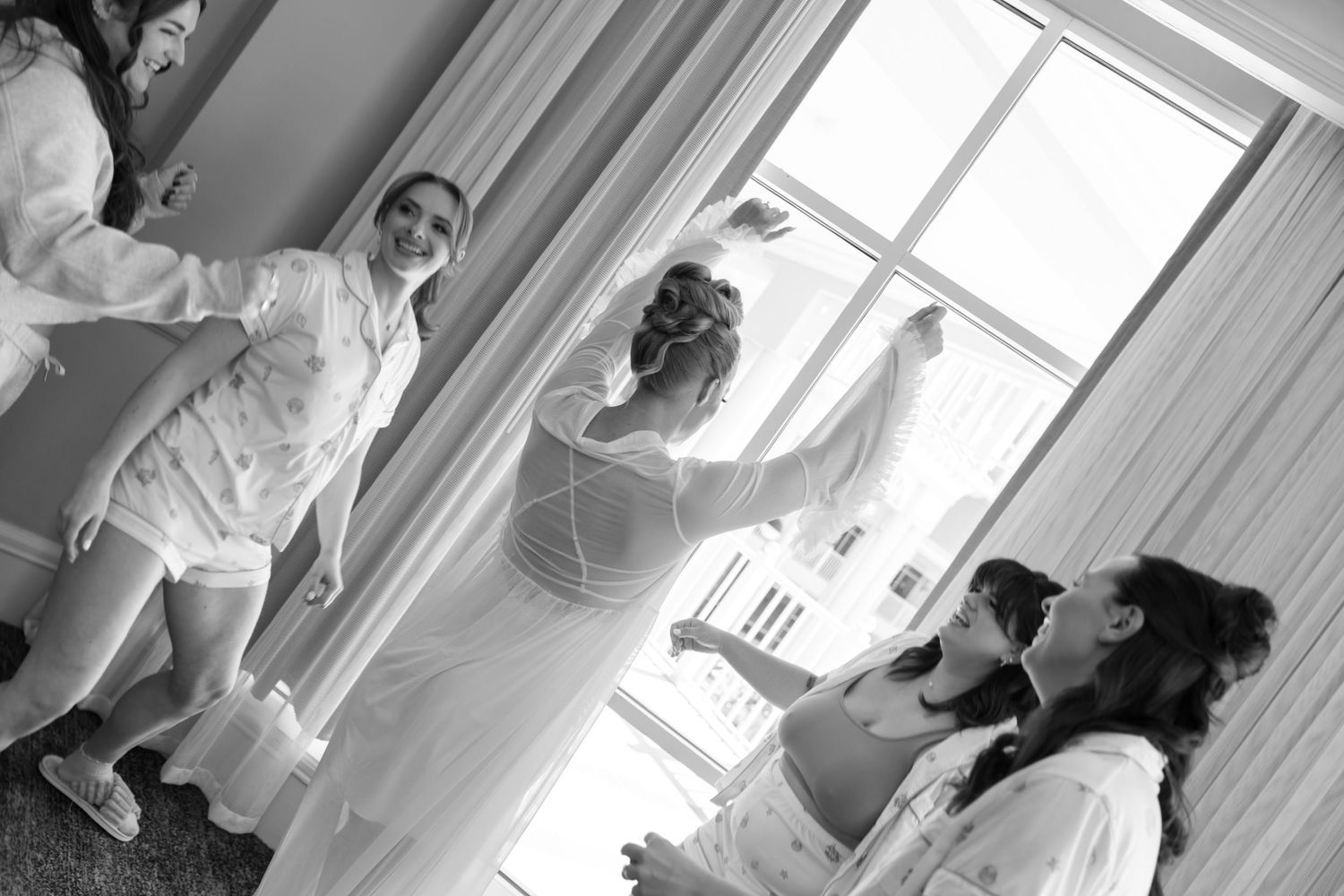 A person in a white dress stands by a window in a bright sunlit room during a bridal preparation moment.