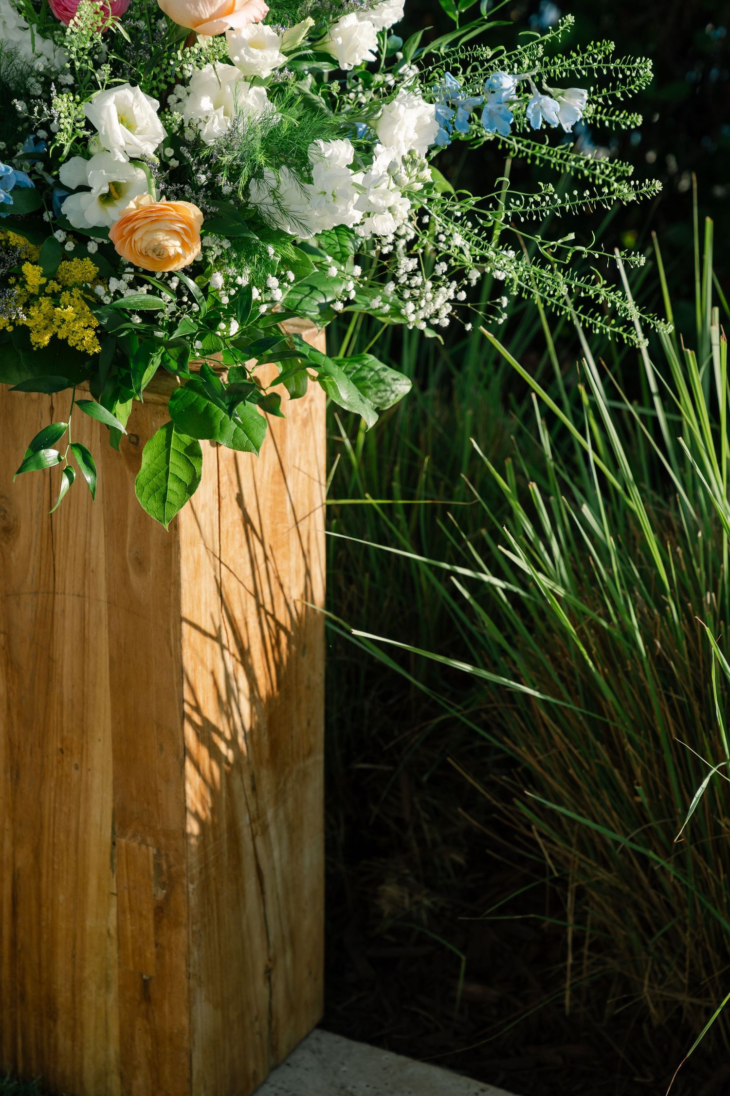 Elegant floral arrangement with white and peach flowers on wooden pedestal against ornamental grass.