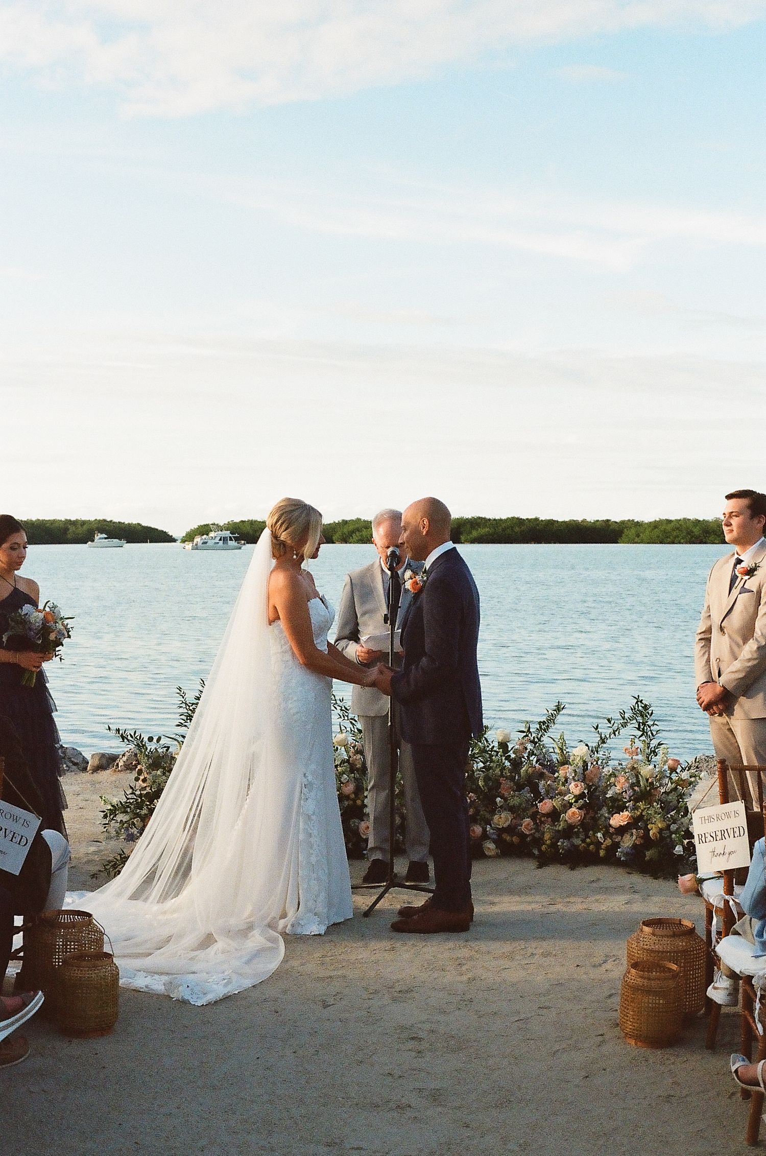 A waterfront wedding ceremony takes place with floral arrangements and rustic wooden decor along the shoreline.