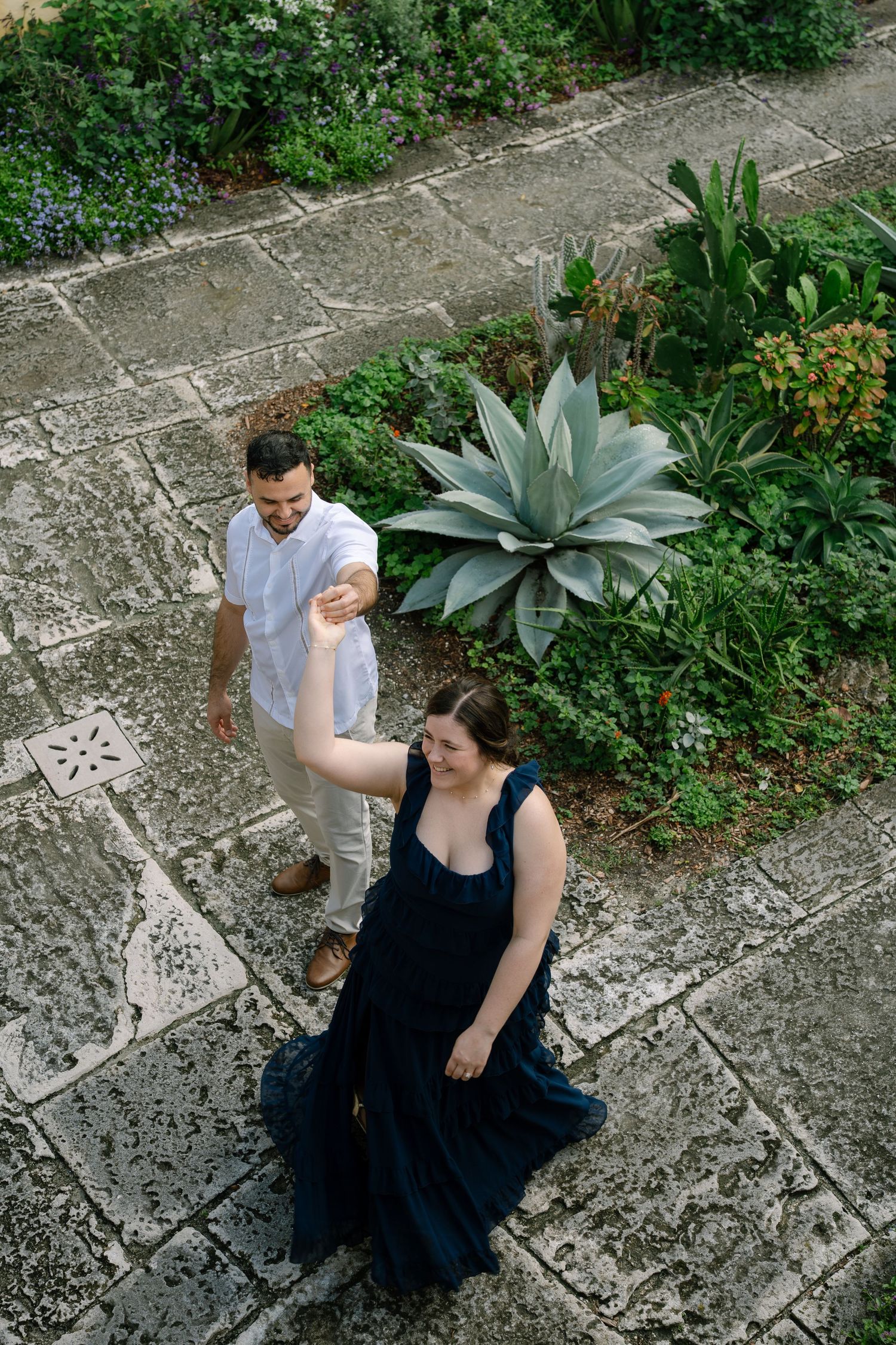 A couple stands on a stone path in a garden with large agave plants and greenery surrounding them.