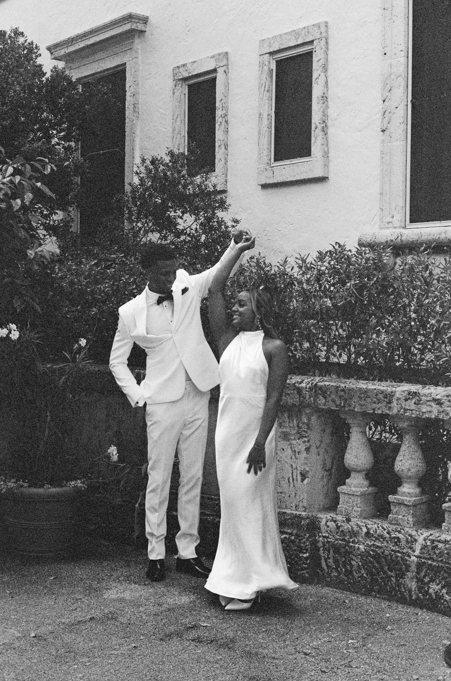 A black and white photo shows a couple in white attire posing on stone steps beside a building with window frames.