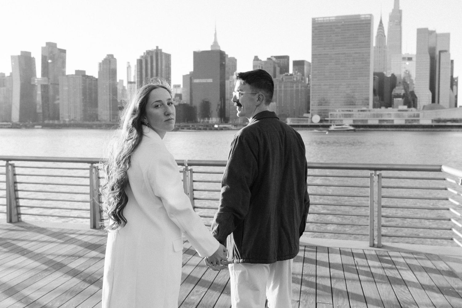A couple poses for romantic photos on a waterfront boardwalk with the Manhattan skyline in the background.