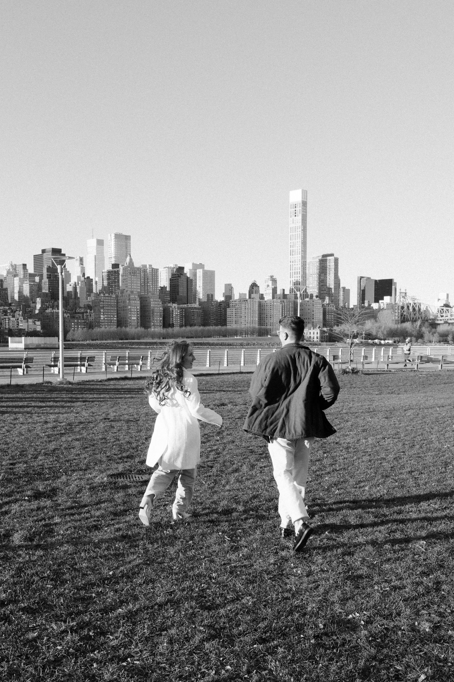 Black and white photo of two figures dancing in a field with Manhattan skyline in background.