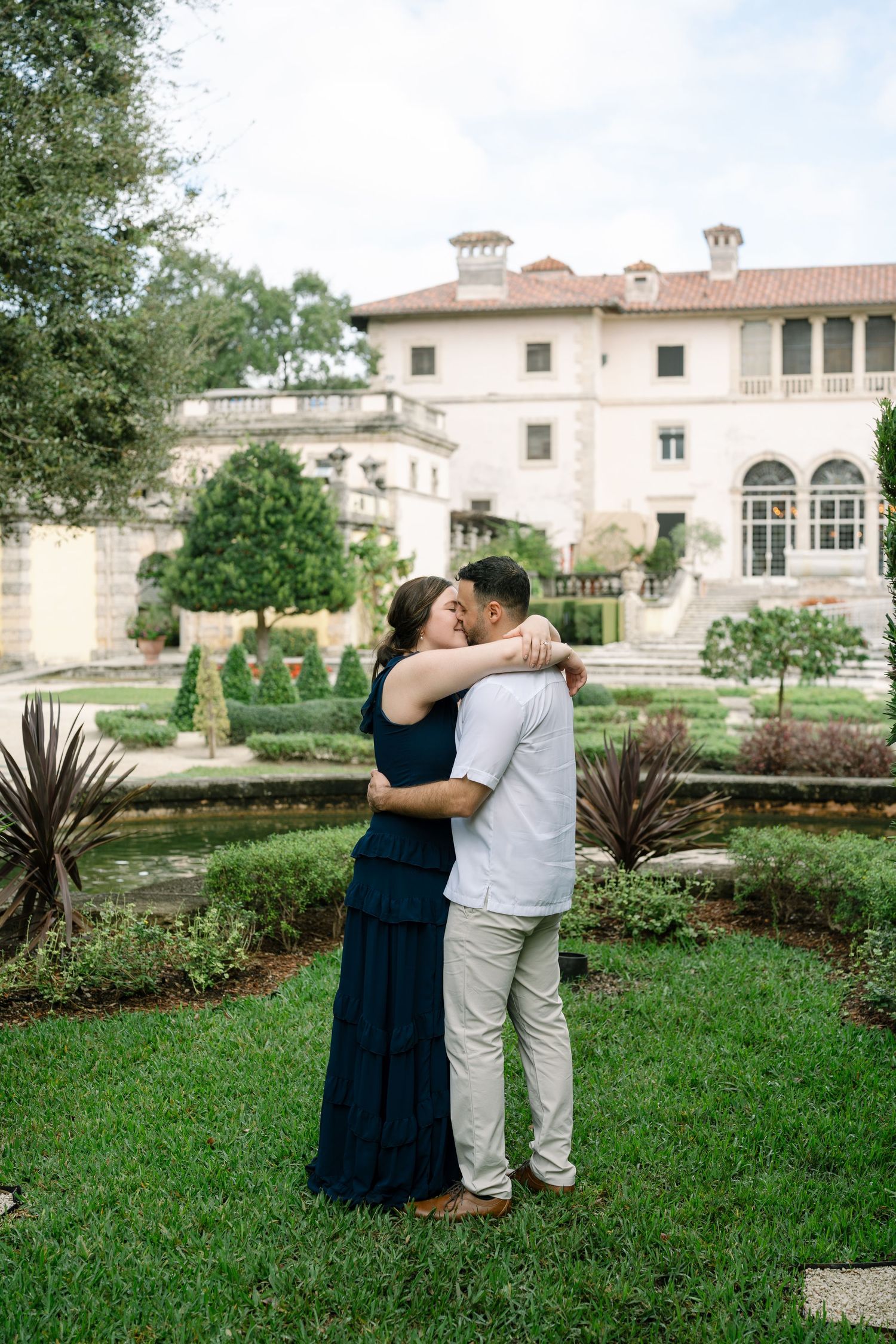 A couple shares an intimate moment in front of an elegant Mediterranean-style mansion with manicured gardens.