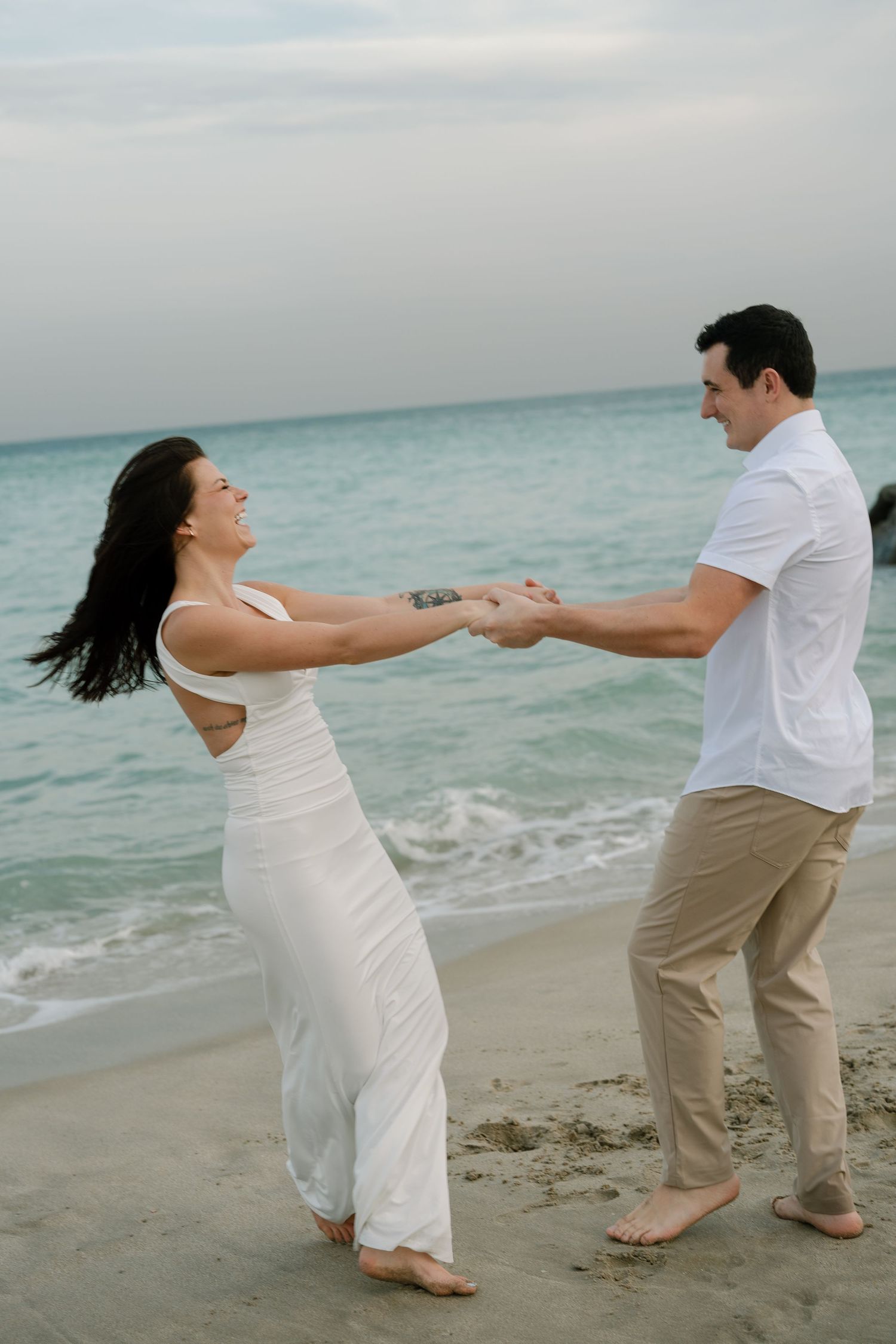 A couple in white and beige clothing dances playfully together on a beach by turquoise waters.